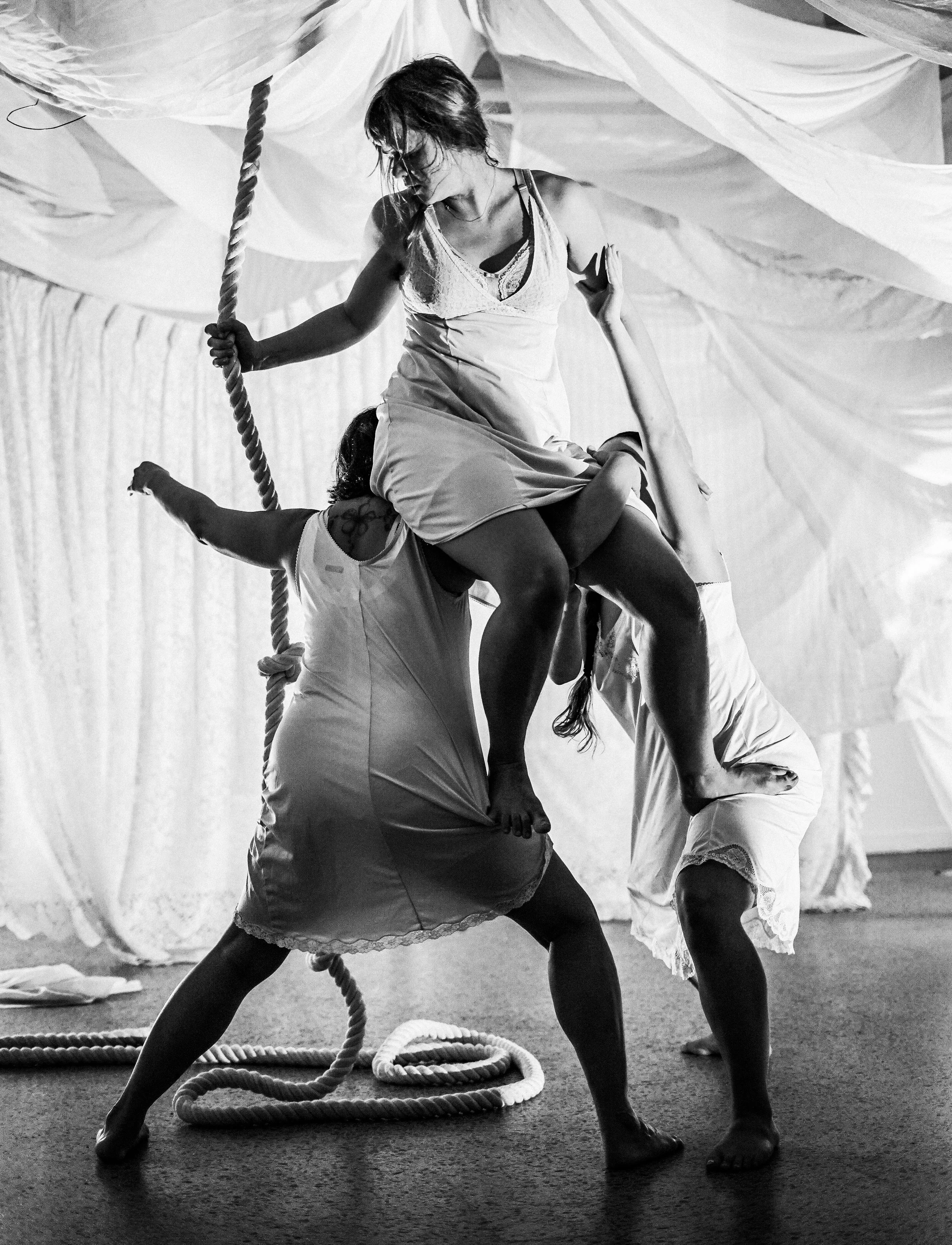 A group of people practicing acro yoga, with one person balancing on another's back while holding onto a rope, in a room with curtains in the background.