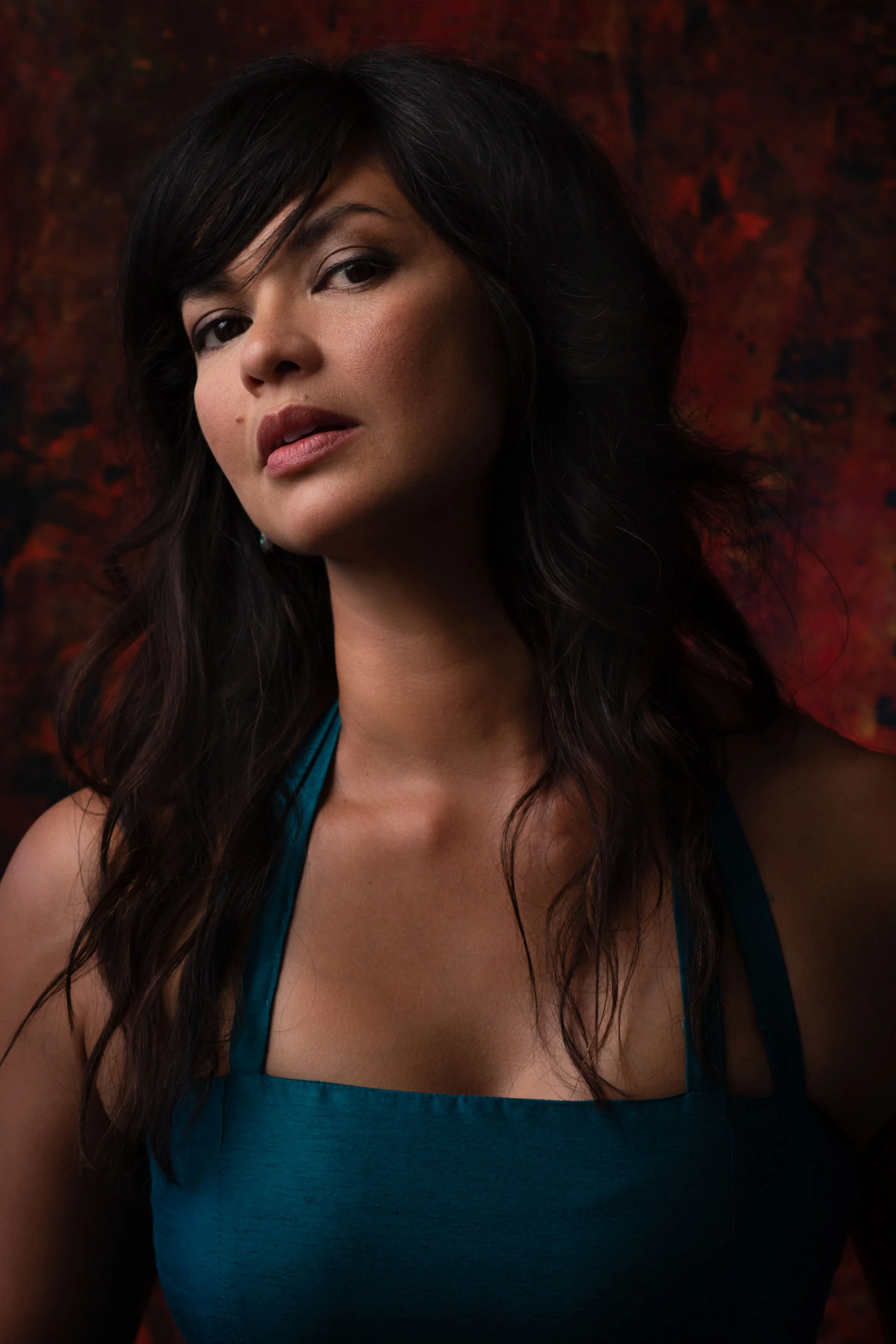 A woman with dark, wavy hair and makeup, wearing a sleeveless teal dress, with a dark and textured red background.