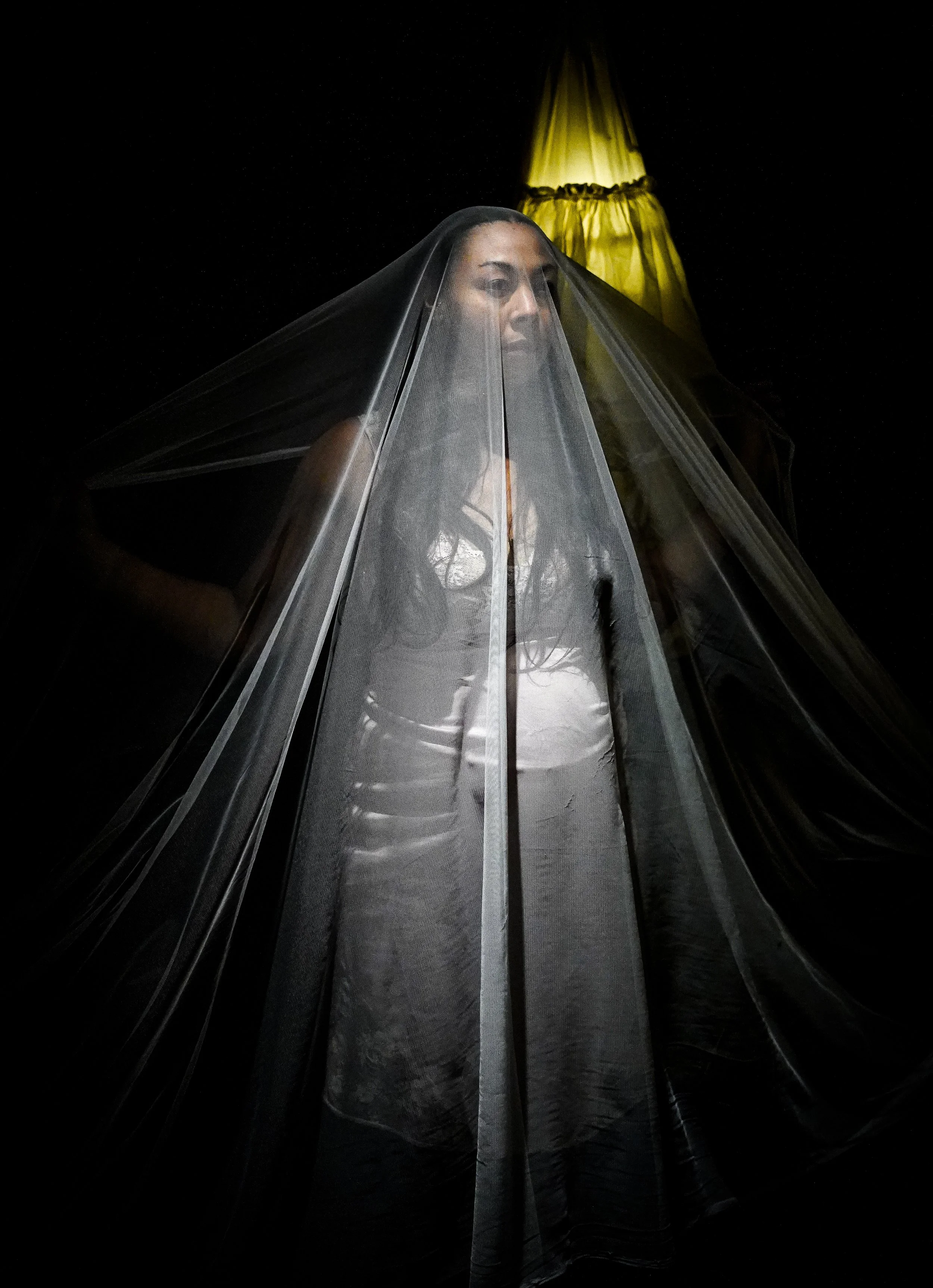A woman with dark hair and facial expression visible is covered by a sheer black veil, standing in front of a yellow lampshade against a dark background.