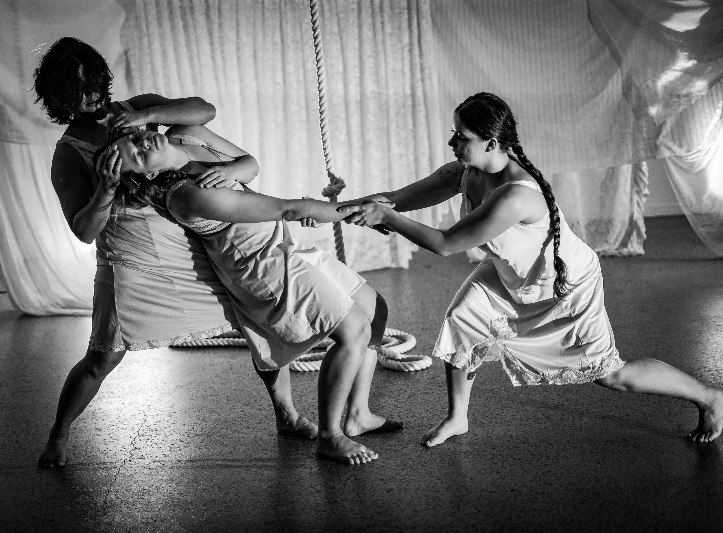Three women are practicing a dance or movement routine together, holding hands and balancing in coordinated poses inside a room with curtains or drapes in the background. The image is black and white.
