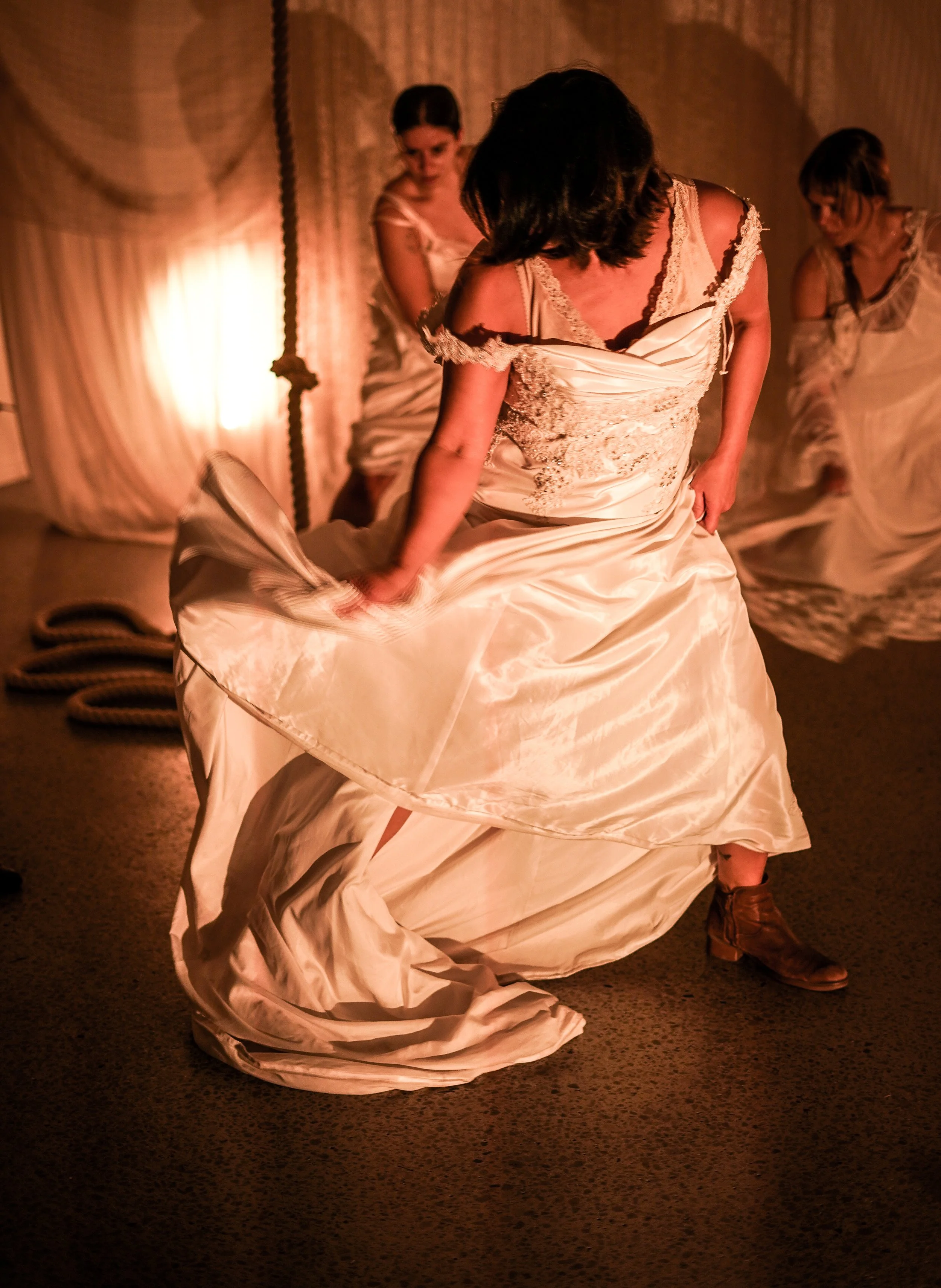 A woman in a white vintage wedding dress, with dark hair, standing in front of a mirror, adjusting her gown. The background includes sheer curtains and two other women in similar dresses, one with glasses and braided hair. The scene is dimly lit with