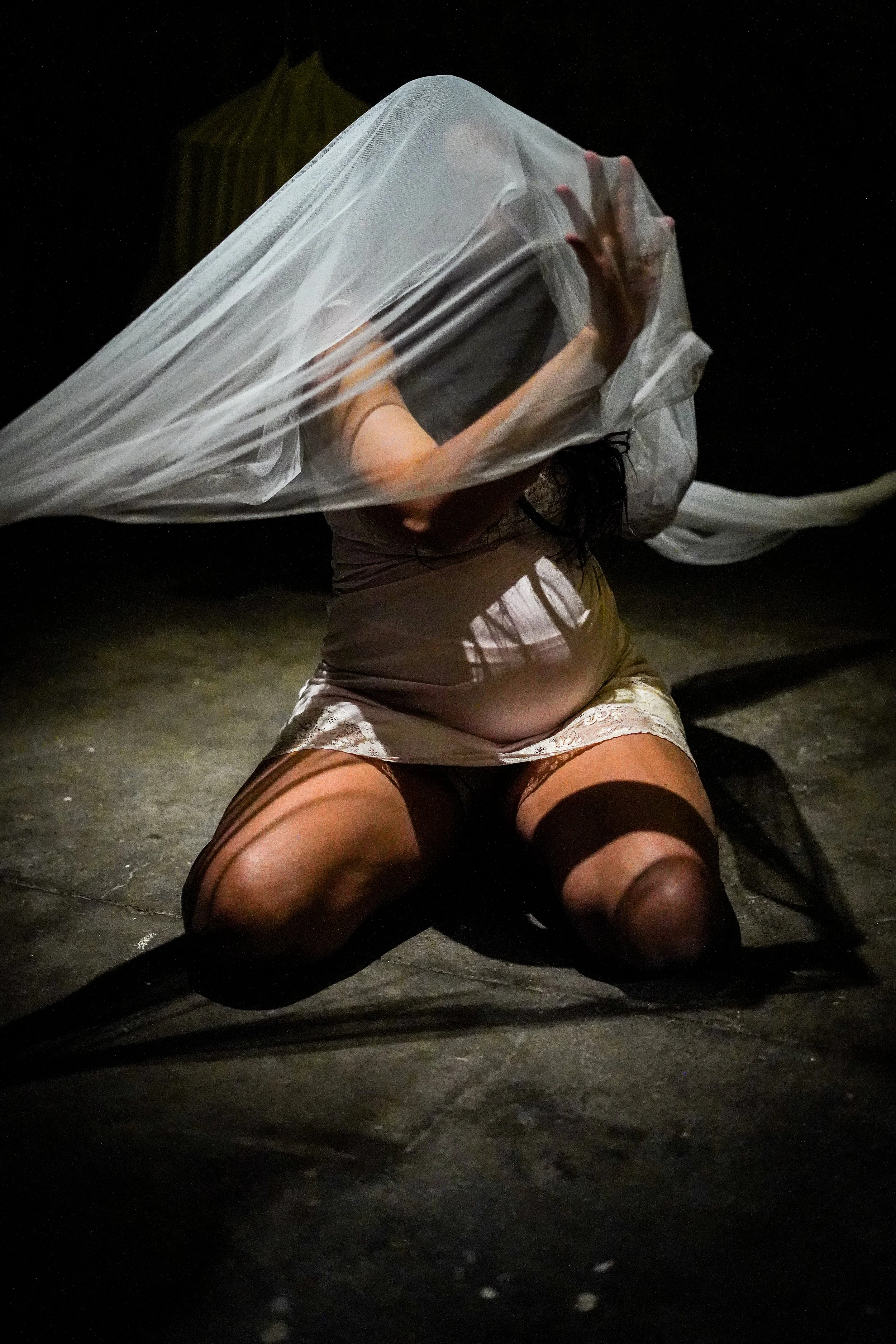 A person is sitting on the ground with their legs spread out and knees bent, wearing a beige skirt and fishnet stockings, with their upper body obscured by a large, transparent plastic sheet covering their head and shoulders. Their right hand is visi