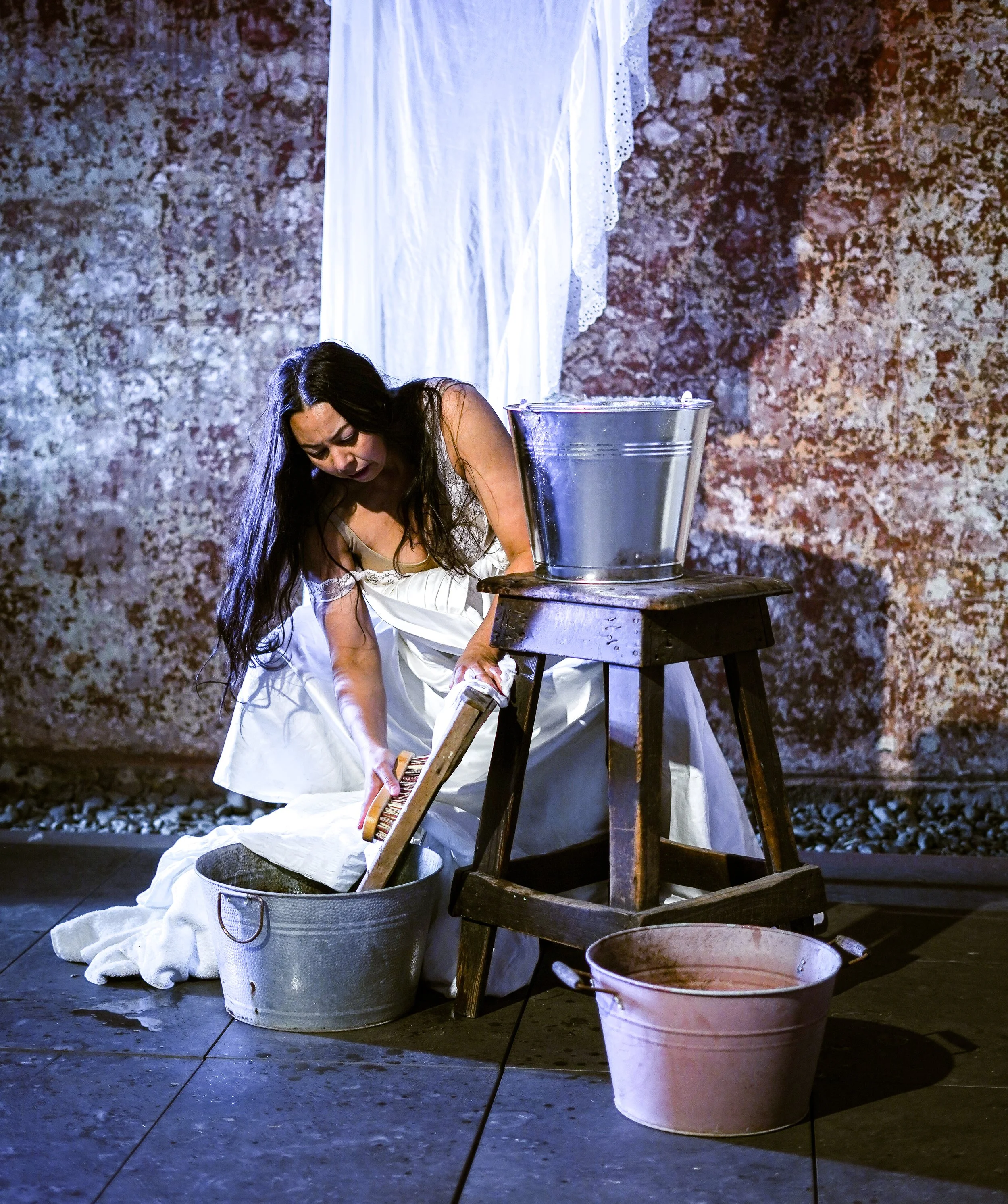 A woman dressed in a white dress is kneeling on the floor, leaning over a large white fabric-covered object, possibly a washing basin. She appears to be washing or cleaning something with a washboard. There is a metal bucket partially visible on the 