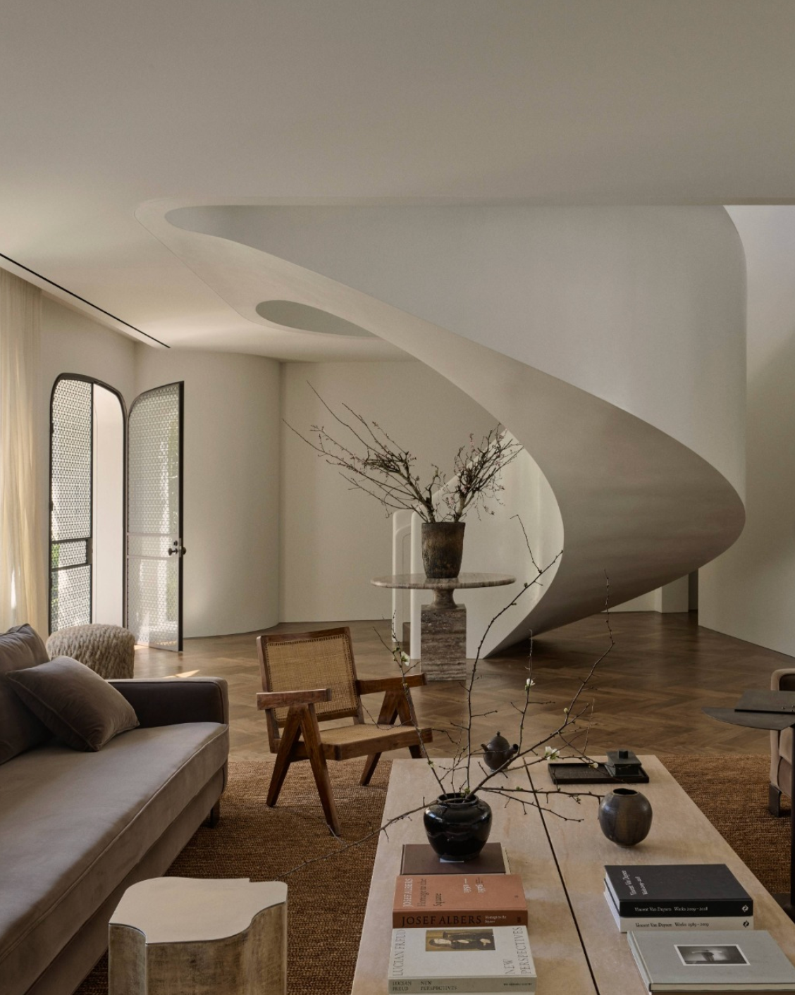 Interior of a modern living room with a staircase, wooden flooring, and a beige sofa. There are decorative items including vases, branches, and books on a white table.