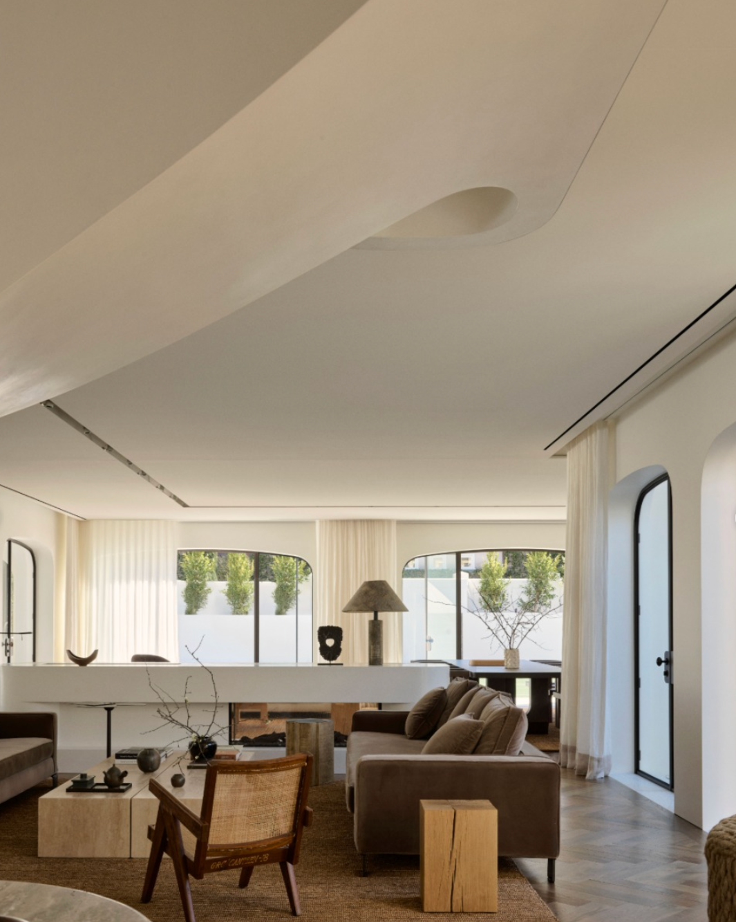 Living room with large windows, beige curtains, and modern furniture including a brown sofa, armchair, and coffee table, decorated with vases and plants.