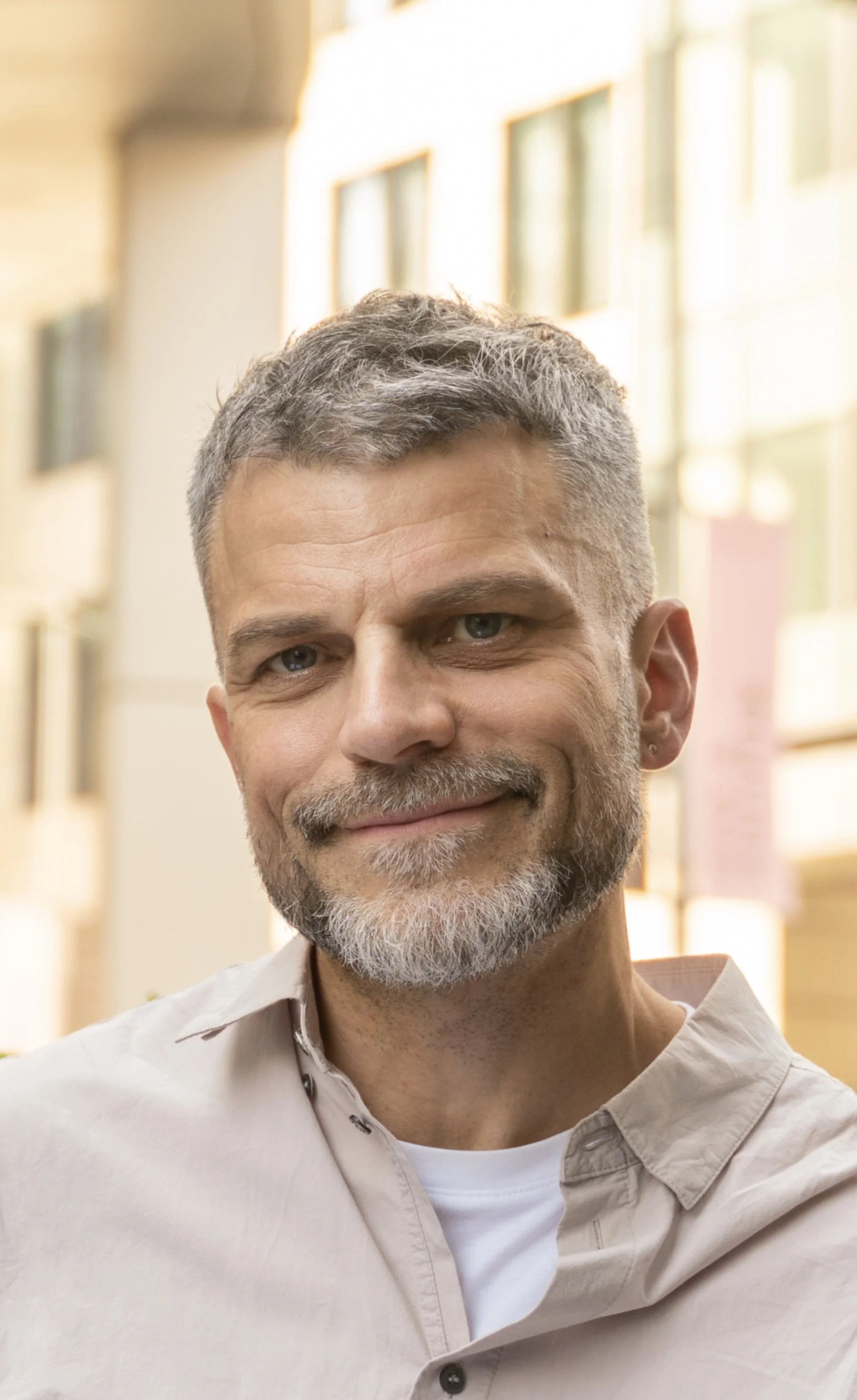Close-up of a smiling middle-aged man with gray hair and beard, wearing a light-colored button-up shirt, standing indoors with large windows in the background.