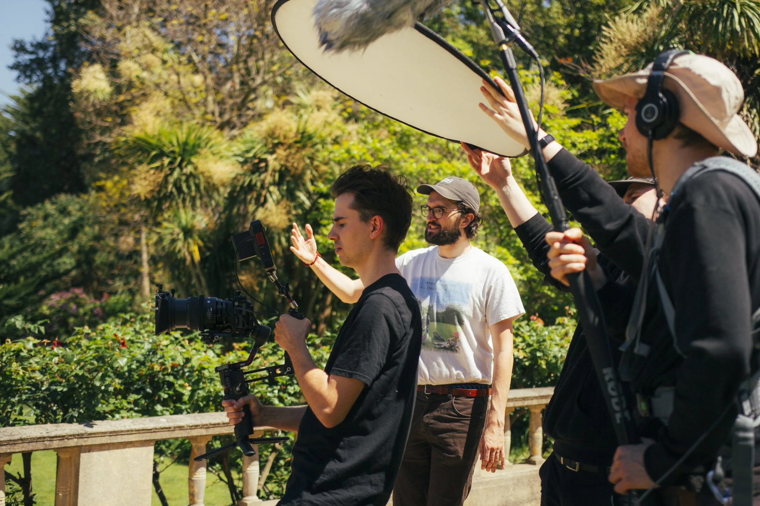 A group of four people working together outdoors on a film or photography project, with lush green trees and bushes in the background. One person is holding a camera with a stabilizer, another is adjusting a light reflector, and the others appear to be involved in the setup or direction, all engaged in a collaborative effort.