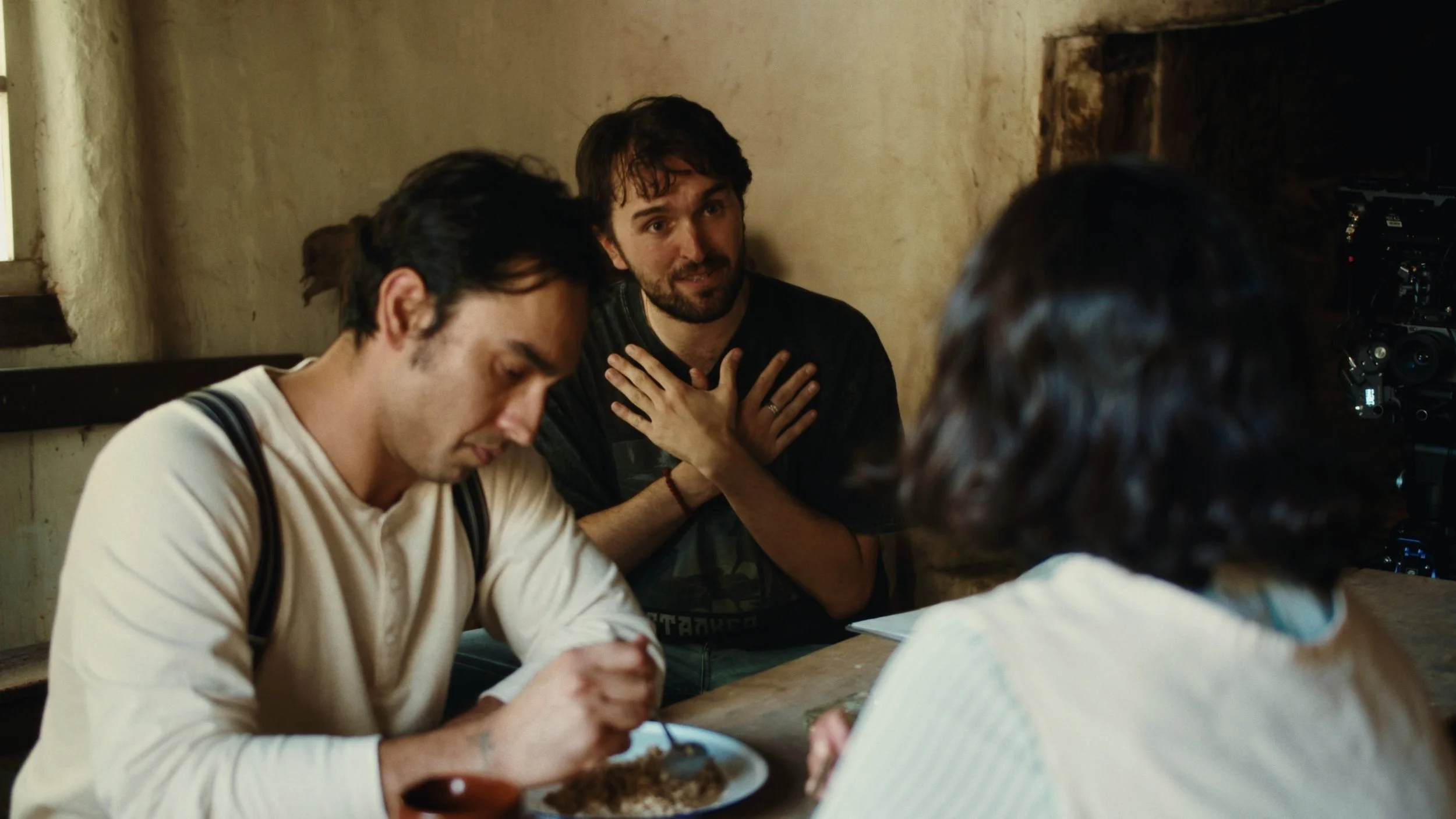 A film crew preparing to shoot a scene inside a rustic, dimly lit room with three people gathered around a table. One person visible in the foreground is eating, while others appear to be discussing or presenting.