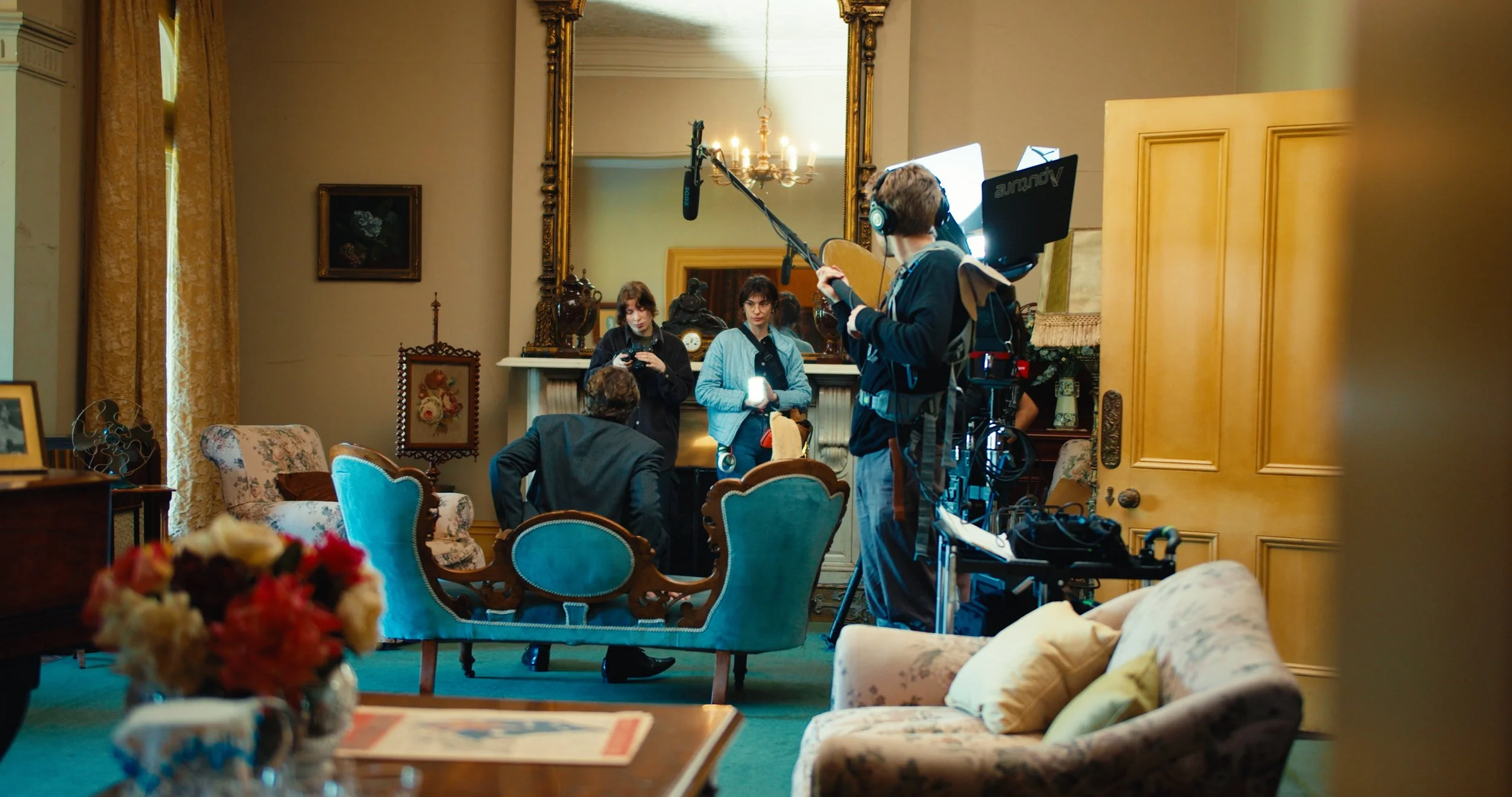 Behind the scenes of a film or television production in a vintage living room with ornate furniture and decorations, with crew members preparing equipment and setting up shots.