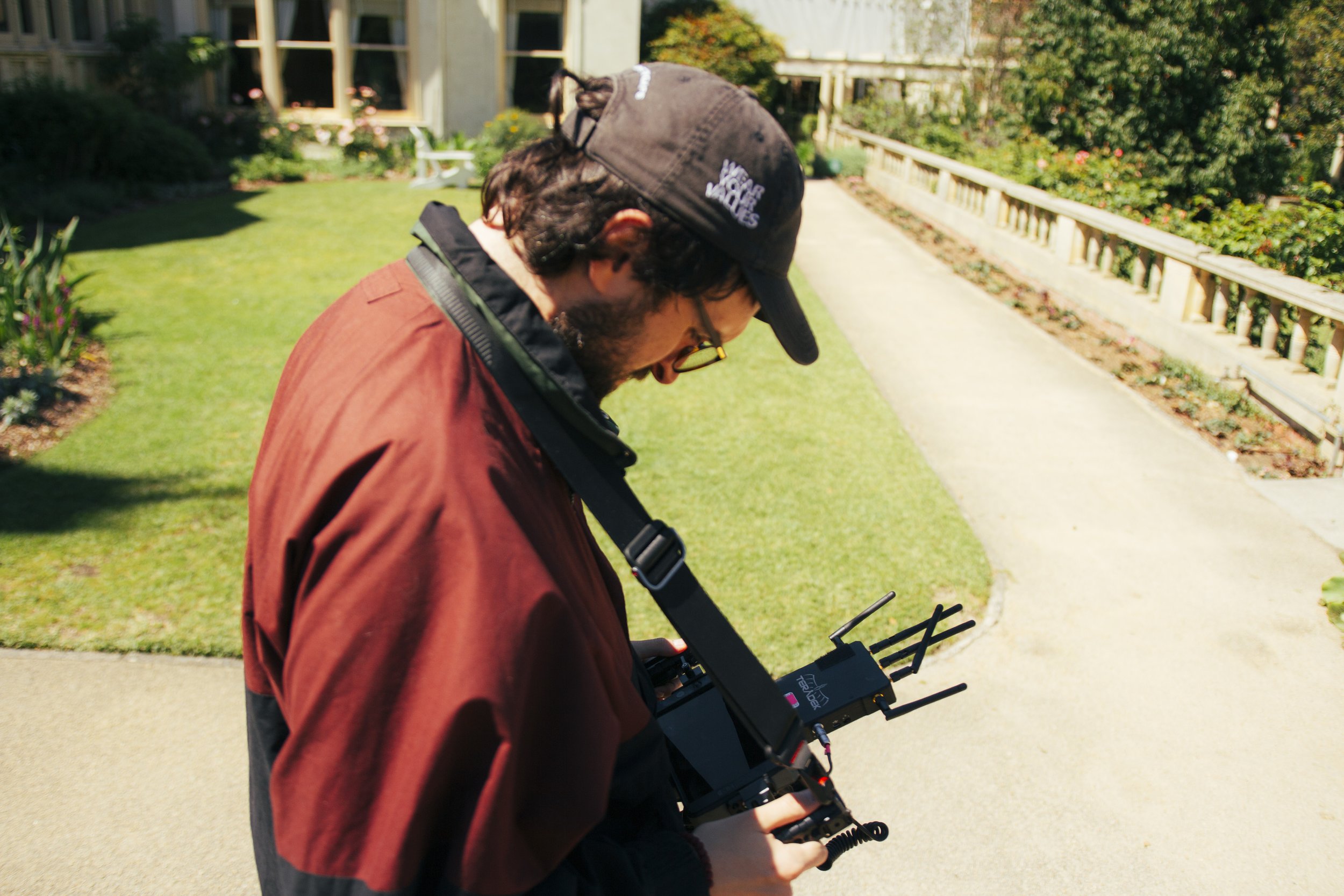 A man wearing a black cap and glasses, dressed in a red jacket, is outdoors on a sidewalk with green lawn and flowering bushes, holding a drone controller and looking at a drone in his hands.