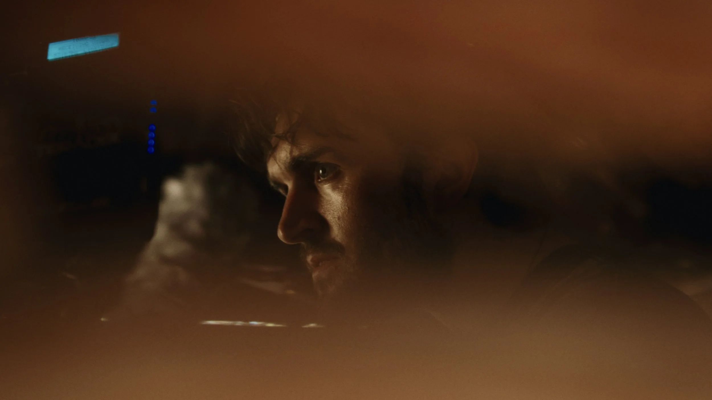 A close-up of a person's face, lying down and looking intently, with a dark background and some blue lights in the distance.