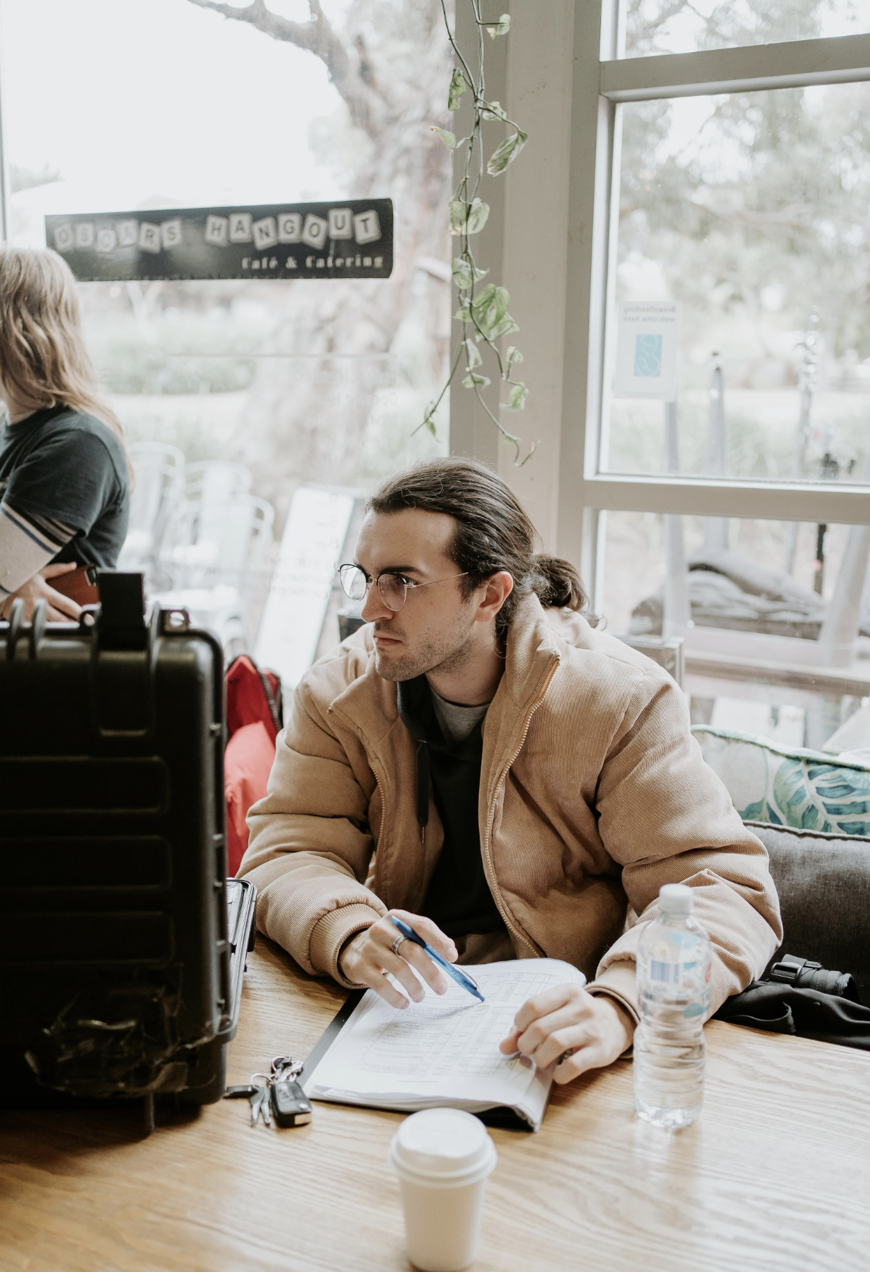 Man with glasses and long hair sitting at a table in a cafe, writing in a notebook with a pen, with a water bottle, coffee cup, and keys on the table, inside a bright cafe with large windows and hanging plants.