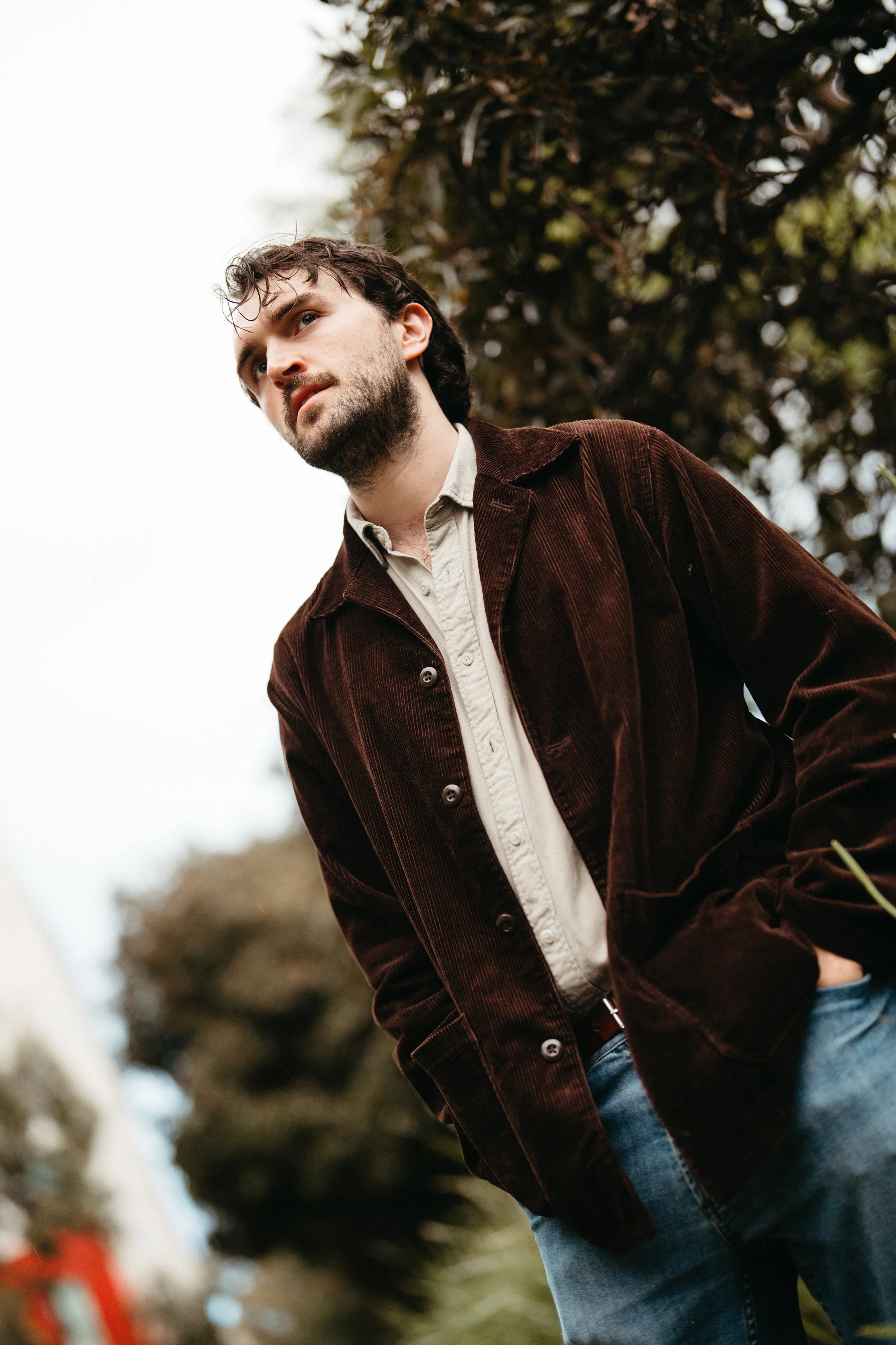 A young man with dark, wavy hair and a beard, wearing a brown corduroy jacket over a light-colored shirt, stands outdoors with a blurred background of trees and overcast sky.