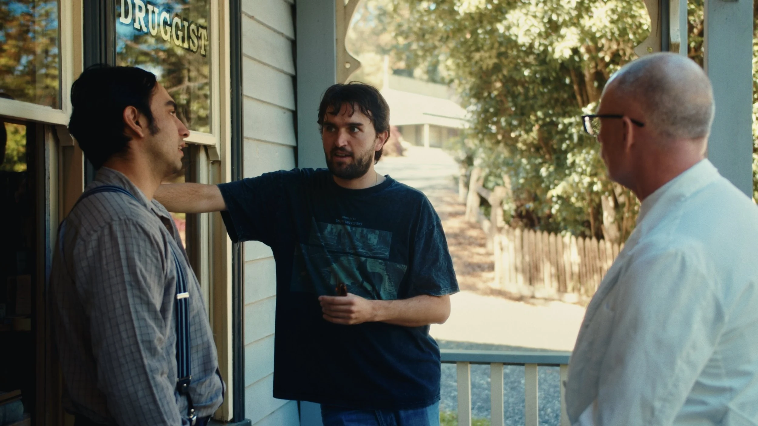 Three men engaged in a conversation outside a building with a sign that reads 'DRUGGIST'. One man, with dark hair and a beard, is in the center and appears to be explaining something. The man to the left has dark hair and is wearing a striped shirt with suspenders, facing the man in the center. The man to the right is bald, wears glasses, and is dressed in a white shirt. The setting suggests a casual outdoor interaction on a sunny day.