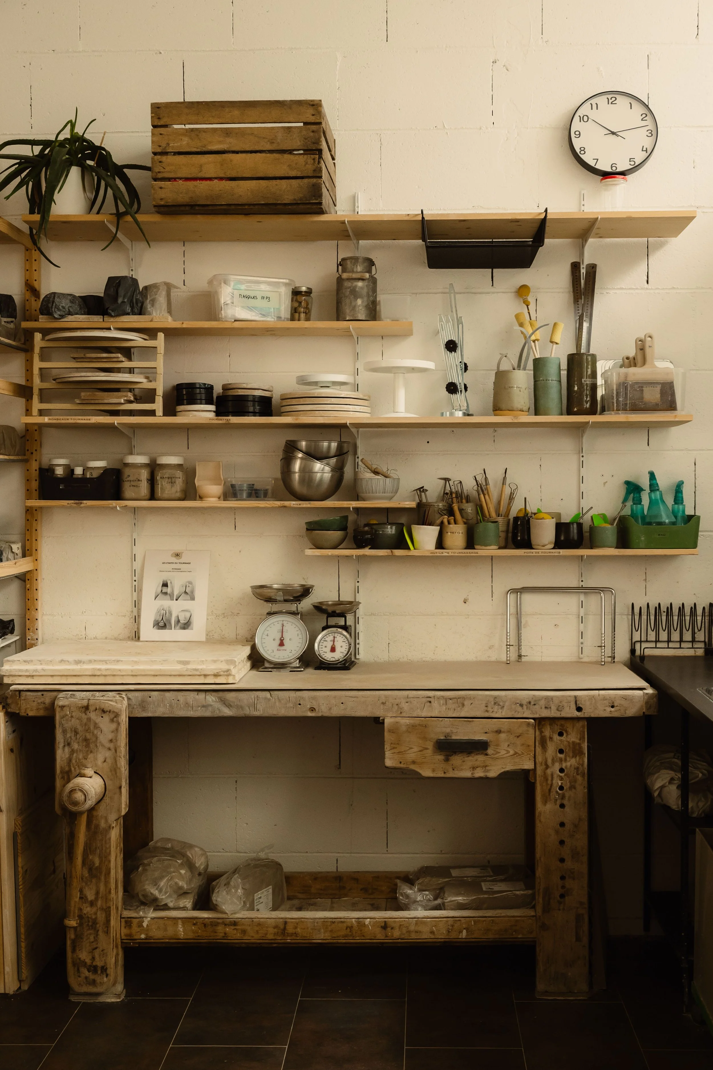 Une étagère en bois fixée à un mur blanc avec des outils de cuisine, des bocaux, des assiettes, et un horloge au mur. Au dessous, un établi en bois avec des sacs en plastique dessous.