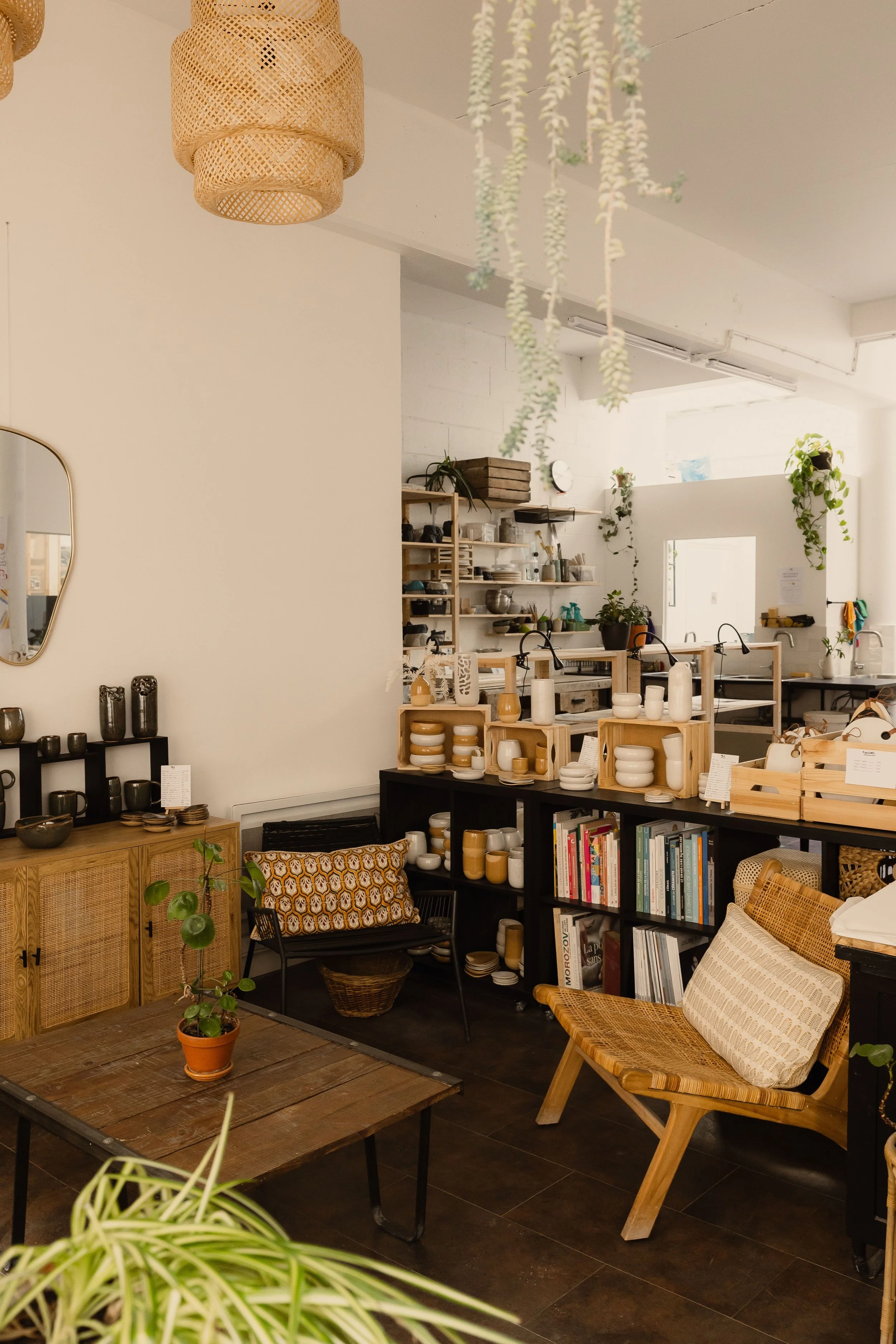 Une boutique ou un espace de décoration intérieure avec des étagères en bois remplies de vaisselle en céramique de couleurs beige et blanc, un canapé en rotin avec coussin, des plantes vertes suspendues et en pot, une table en bois avec une plante en