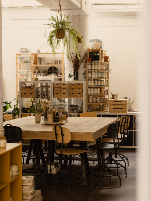 Intérieur d'une atelier de création ou d'une cuisine, avec une grande table en bois entourée de chaises, étagères avec divers contenants et plantes décoratives.