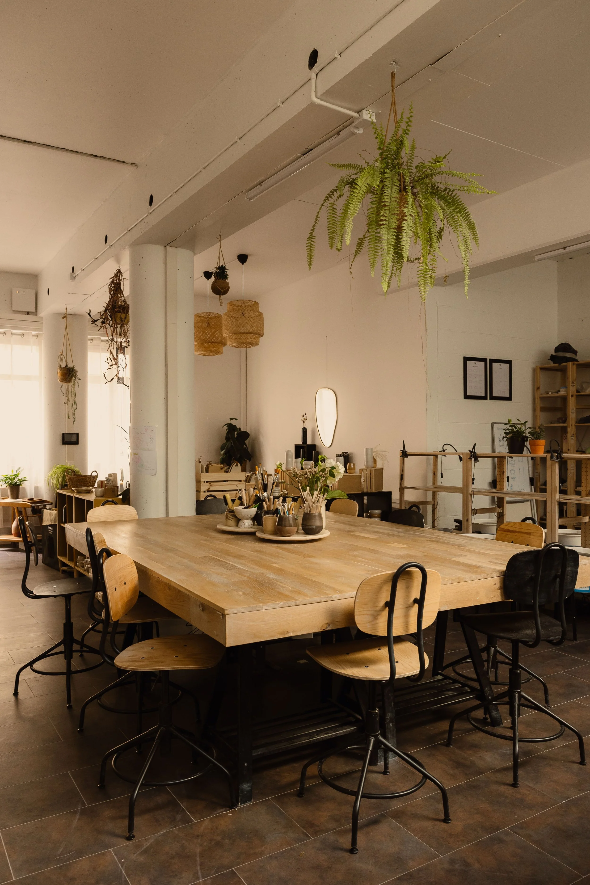 Une salle à manger ou un espace de travail avec une grande table en bois entourée de chaises, decorée de vases et d'objets de bureau, avec des plantes suspendues au plafond, des lampes en rotin et un mur blanc avec cadres et étagères.