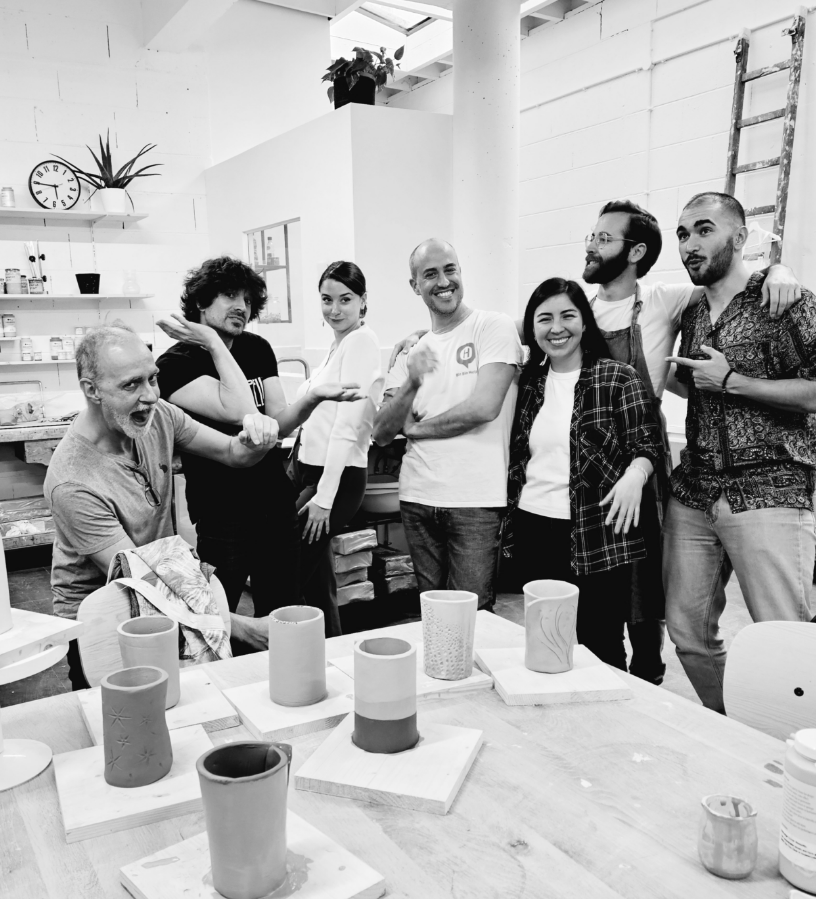Un groupe de sept personnes debout dans un atelier, entouré de céramiques en cours de création. Certains sourient, d'autres prennent une pose décontractée. Le fond montre des étagères avec des fournitures d'art et un escalier en bois.