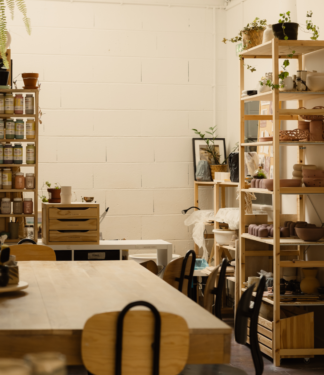 Une pièce intérieure avec des étagères en bois contenant des pots de plantes, des céramiques et des fournitures diverses, un grand tableau à l'arrière, et une table en bois avec des chaises.