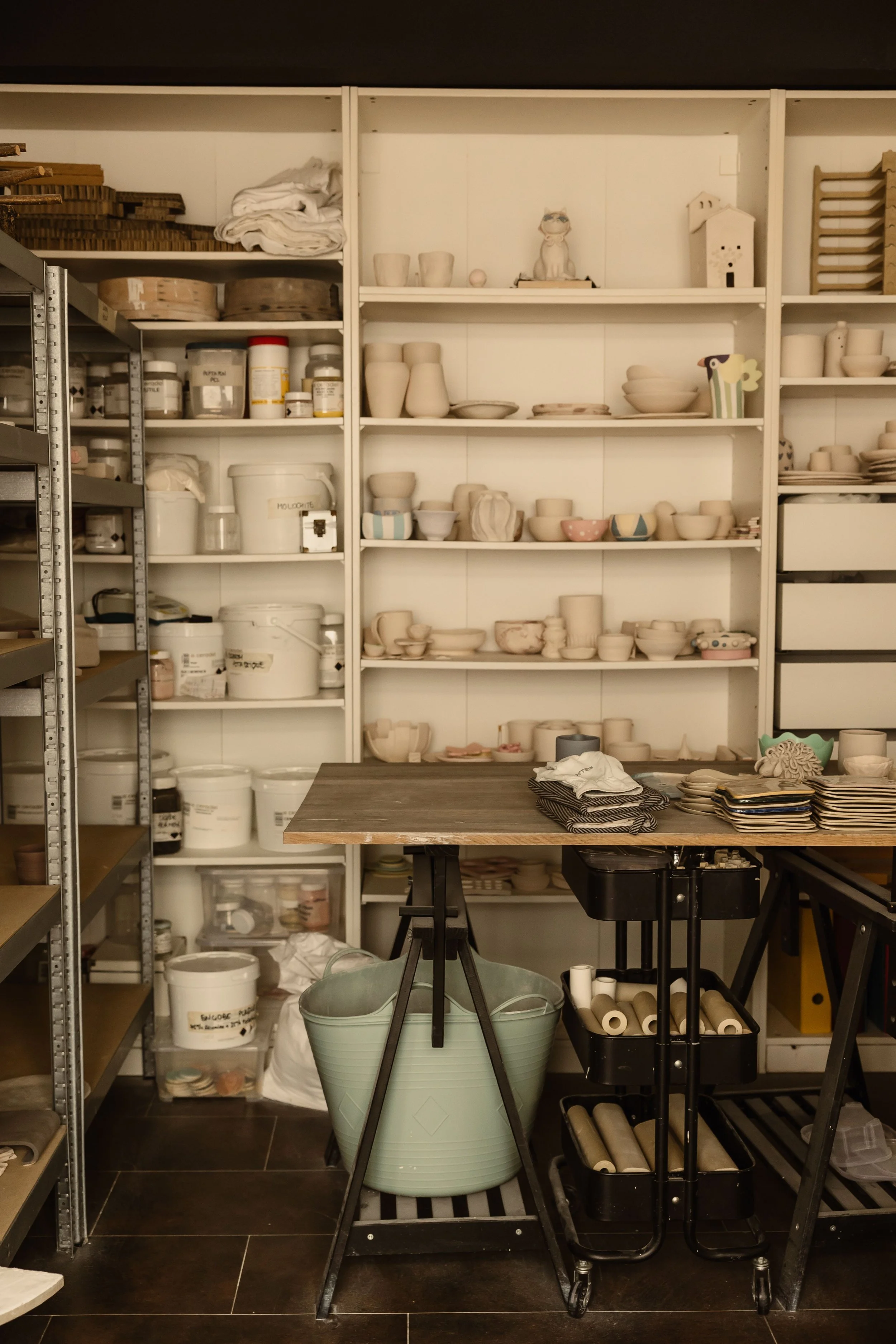 Etagère remplie de matériel de poterie, y compris des pots, des pâtes, des rouleaux, et des outils pour la fabrication d'objets en céramique, avec une table en bois au centre.