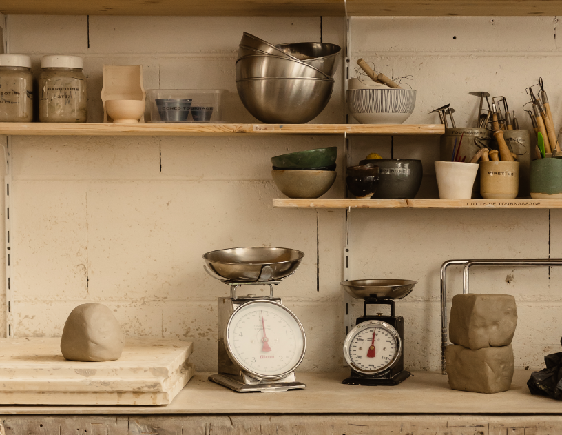 Étagère avec divers ustensiles de cuisine, deux balances, et des pierres pour la poterie sur un plan de travail