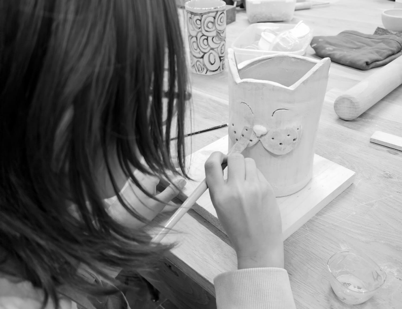 Une personne en train de peindre une poterie en céramique représentant un visage de chat avec un pinceau, assise à une table avec divers outils et matériaux de sculpture.