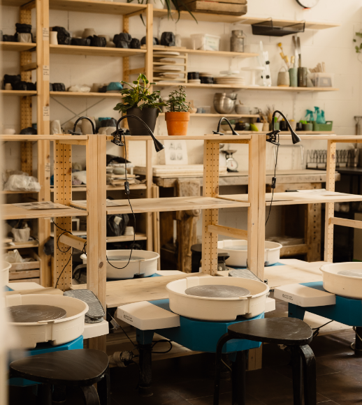 Atelier de poterie équipé de tours de potier, étagères avec matériel et plantes.