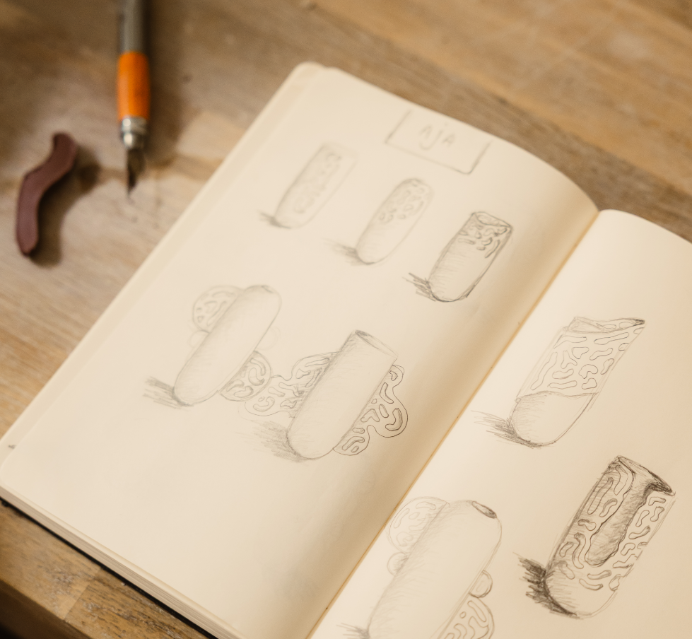 Sketchs de cylindres avec motifs et nuances de dessin sur une page d'un carnet, avec un crayon, une gomme et un cutter à côté.
