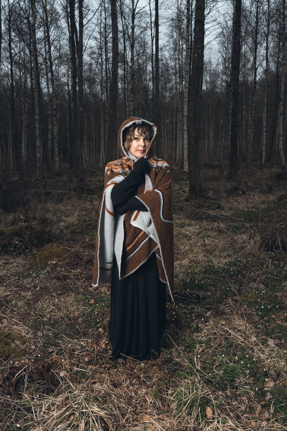 En kvinna står i en skog under dagtid, iklädd en brun och grå poncho med luva och svart klänning, med en stav eller pinne i handen.
