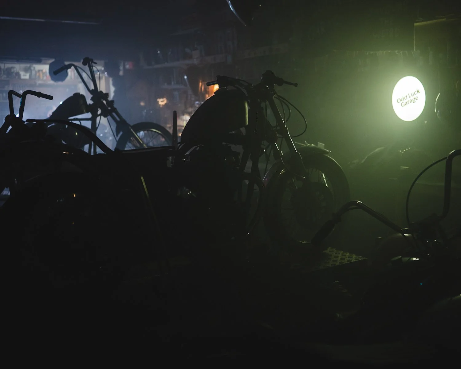 Mörk bild av motorcyklar i ett garage med svagt ljus, en rund ljusskylt med texten 'Odd Luck Garage' syns på bakgrunden.