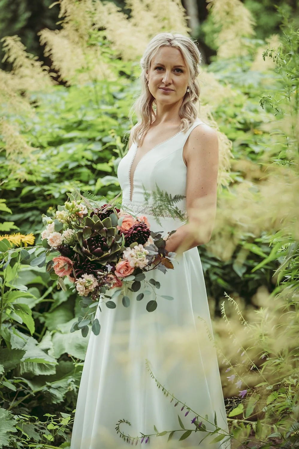 Ung kvinna i bröllopsklänning håller en stor blomsterbukett i en lummig trädgård.