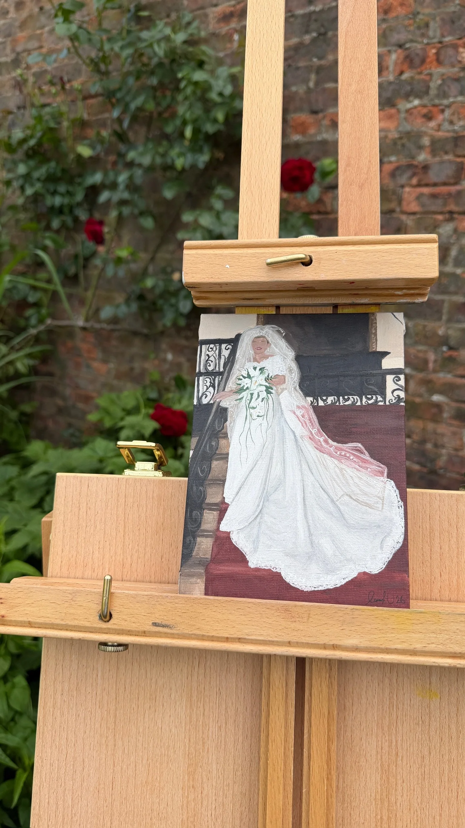 A painting of a bride in a white wedding dress holding a bouquet, displayed on an easel outdoors with a brick wall and red roses in the background.