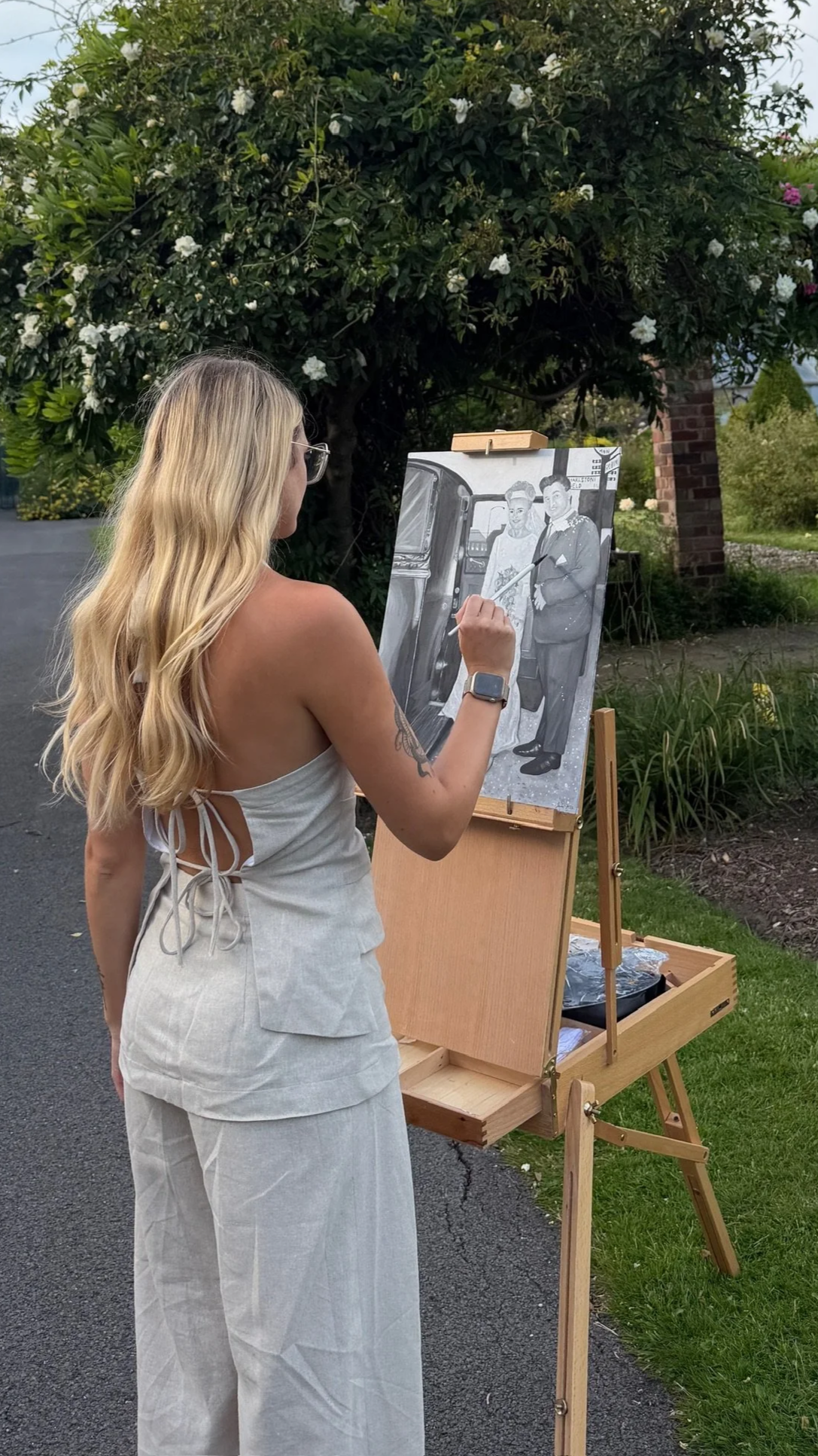 A woman with long blonde hair, wearing glasses and a light-colored sleeveless dress, is painting on a canvas outdoors. The painting depicts two people, a woman and a man, standing in front of a vintage car, with the woman wearing a dress and the man in a suit. She is using a brush to add details to the artwork, which is mounted on an easel.