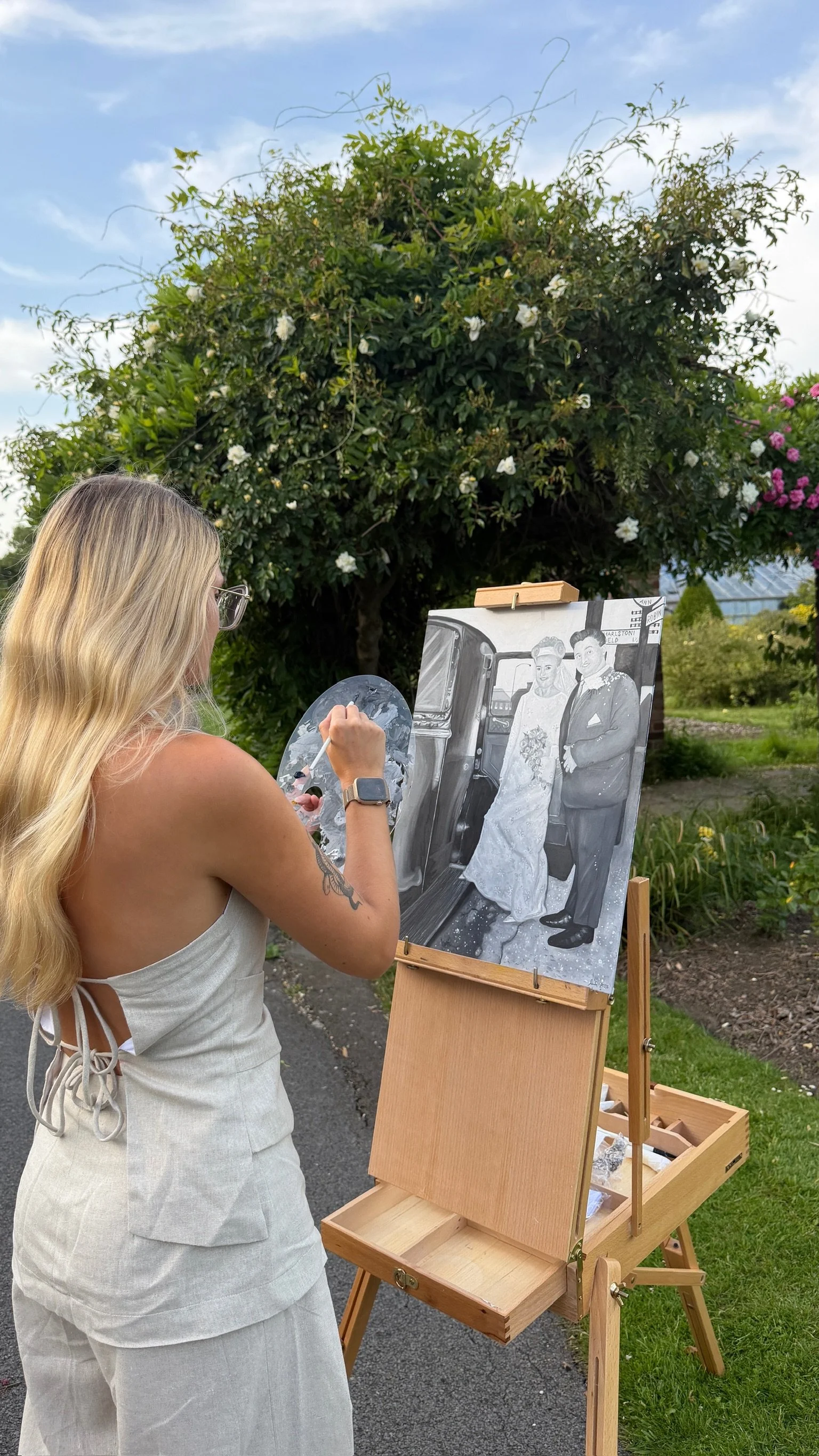 An artist painting a black and white portrait of a bride and groom on a large canvas outdoors. The artist is a woman with long blonde hair, wearing glasses and a sleeveless white dress, standing next to an easel with painting supplies. The background has greenery, flowering bushes, and a blue sky.