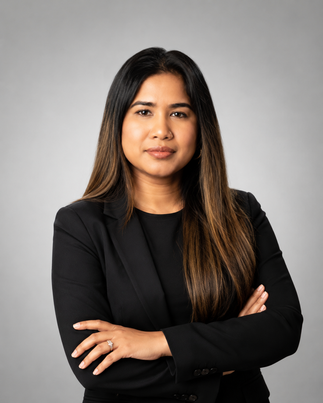 A woman with long dark hair, wearing a black blazer, standing with arms crossed against a plain gray background.