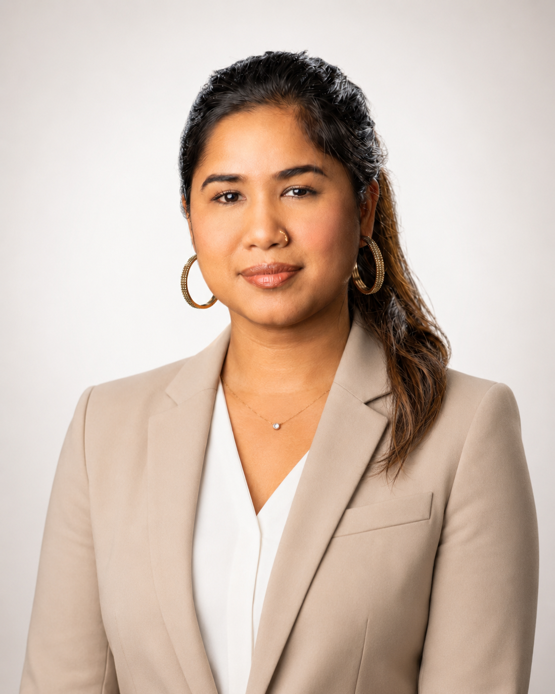Professional woman with dark hair, wearing a beige blazer, white blouse, hoop earrings, and a small necklace, posing against a plain light background.
