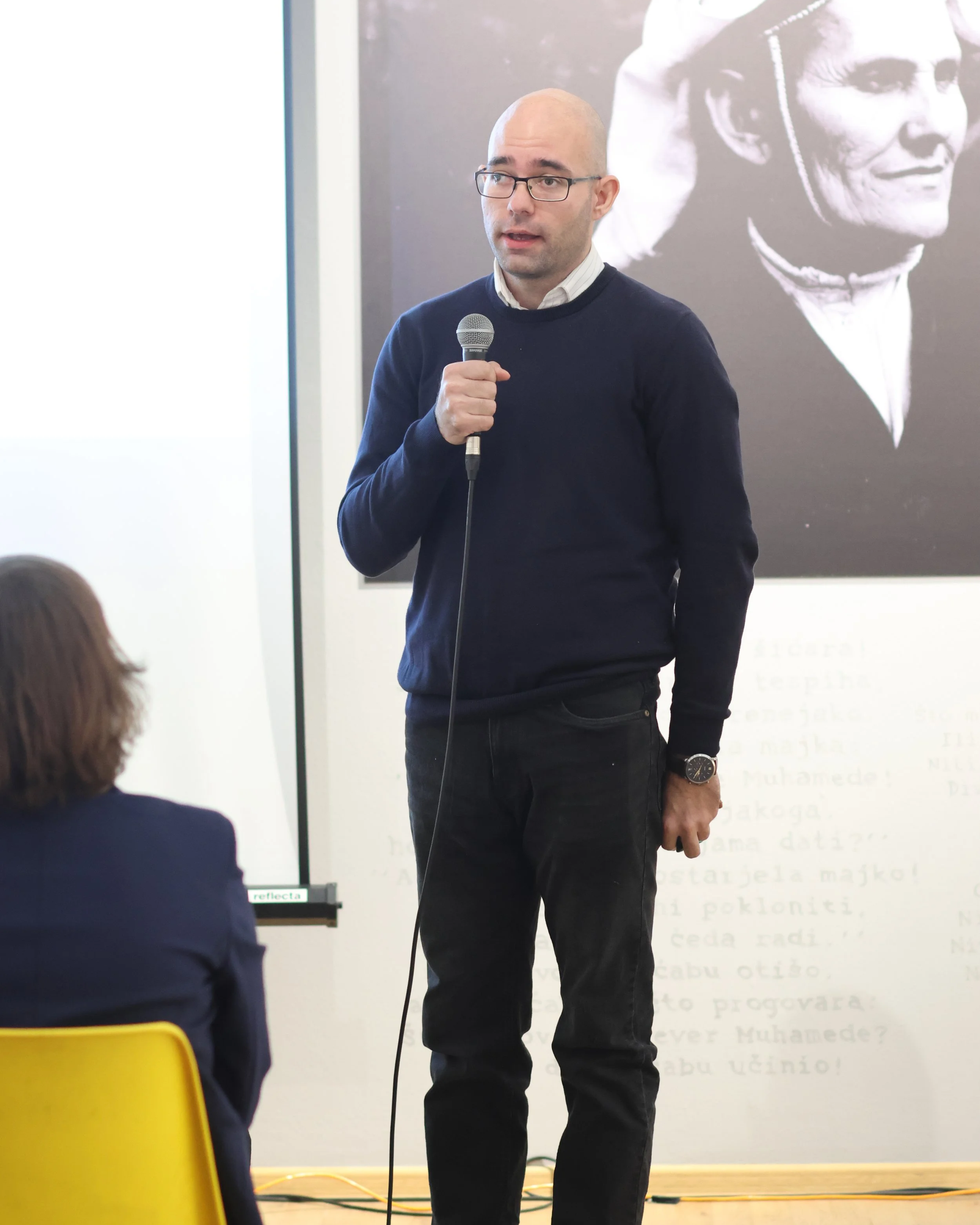 A man with glasses and a watch, wearing a dark sweater over a light collared shirt, is holding a microphone and speaking. In the foreground, someone with a dark blazer is visible, sitting and facing the man. The setting appears to be a presentation or lecture in a classroom or conference room.