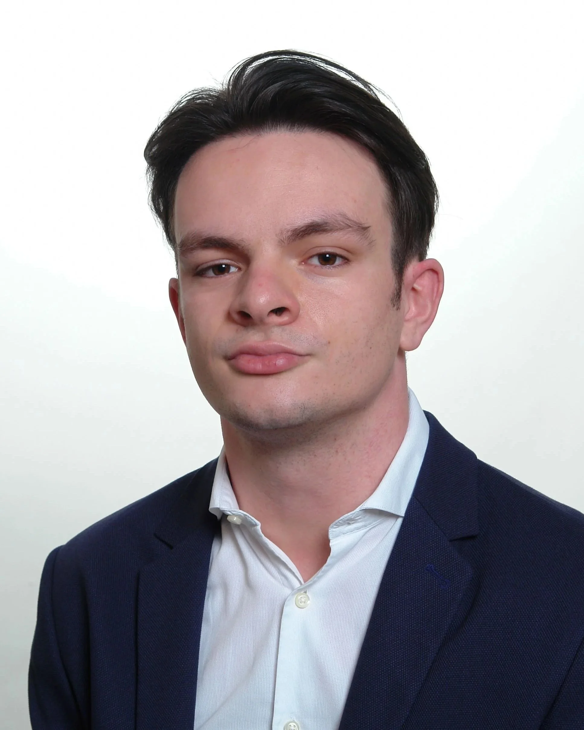 Close-up of a young man's face and neck, wearing a white shirt and navy blazer, against a plain white background.