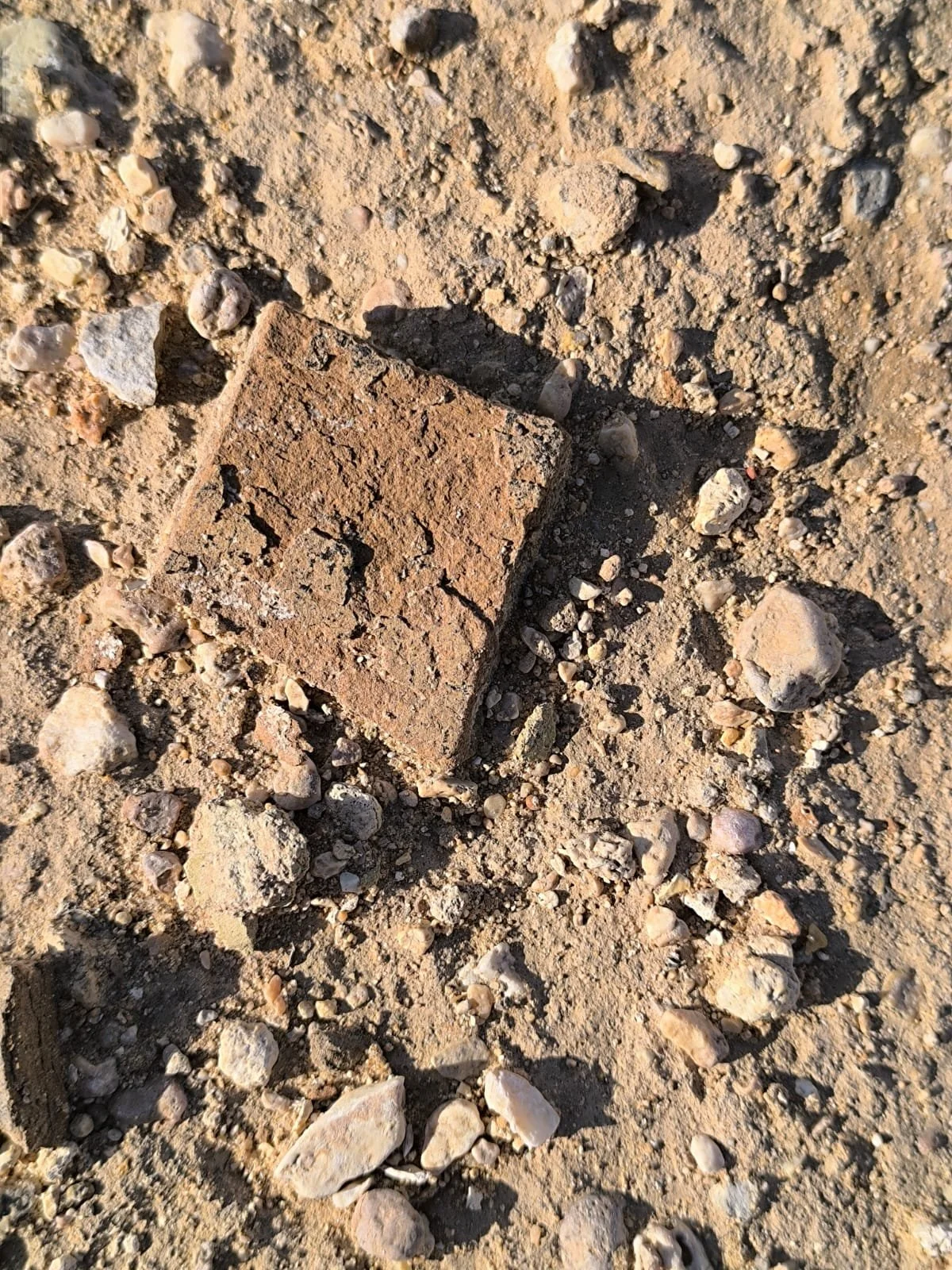 A close-up of a pottery fragment lying on a dirt and gravel surface with small rocks and pebbles scattered around.