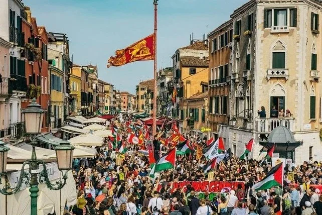 Crowd of people holding Palestinian flags during a demonstration in a busy city street lined with colorful buildings.