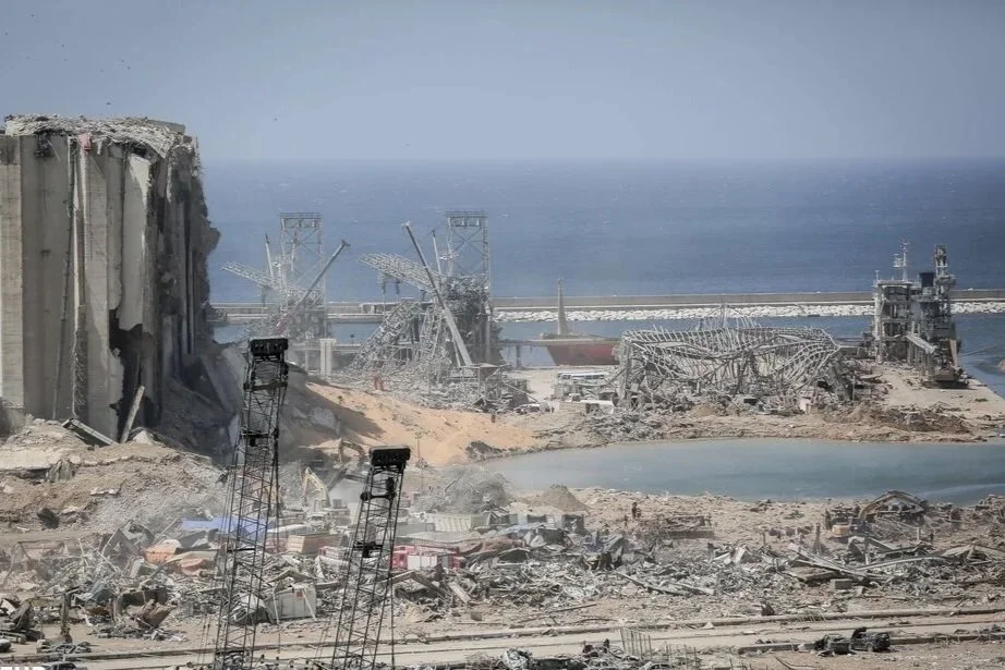 Damaged and collapsed buildings in a coastal city, with debris scattered on the ground and the ocean in the background.