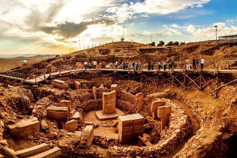 Archaeological excavation site with ancient structures, wooden walkways, and people exploring, under a partly cloudy sky.