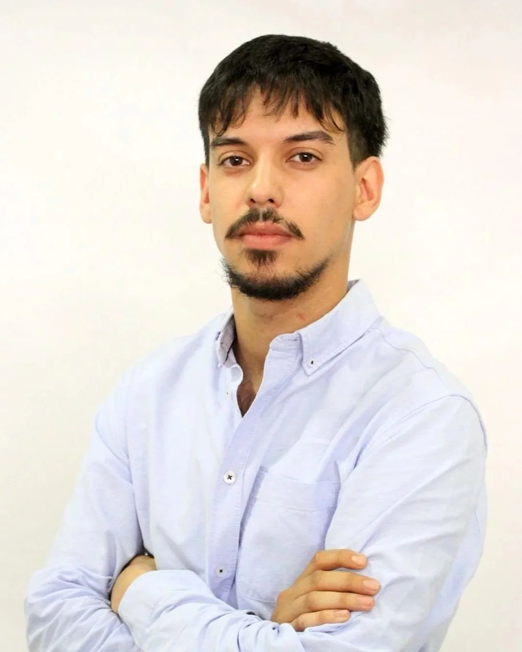 A young man with dark hair, facial hair, and light skin wearing a light-colored button-up shirt standing against a plain white background.