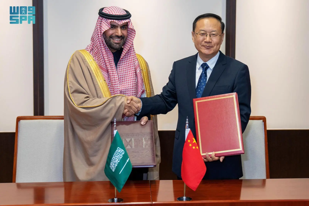 Two men shaking hands at a formal signing ceremony, with Saudi and Chinese flags on the table in front of them.