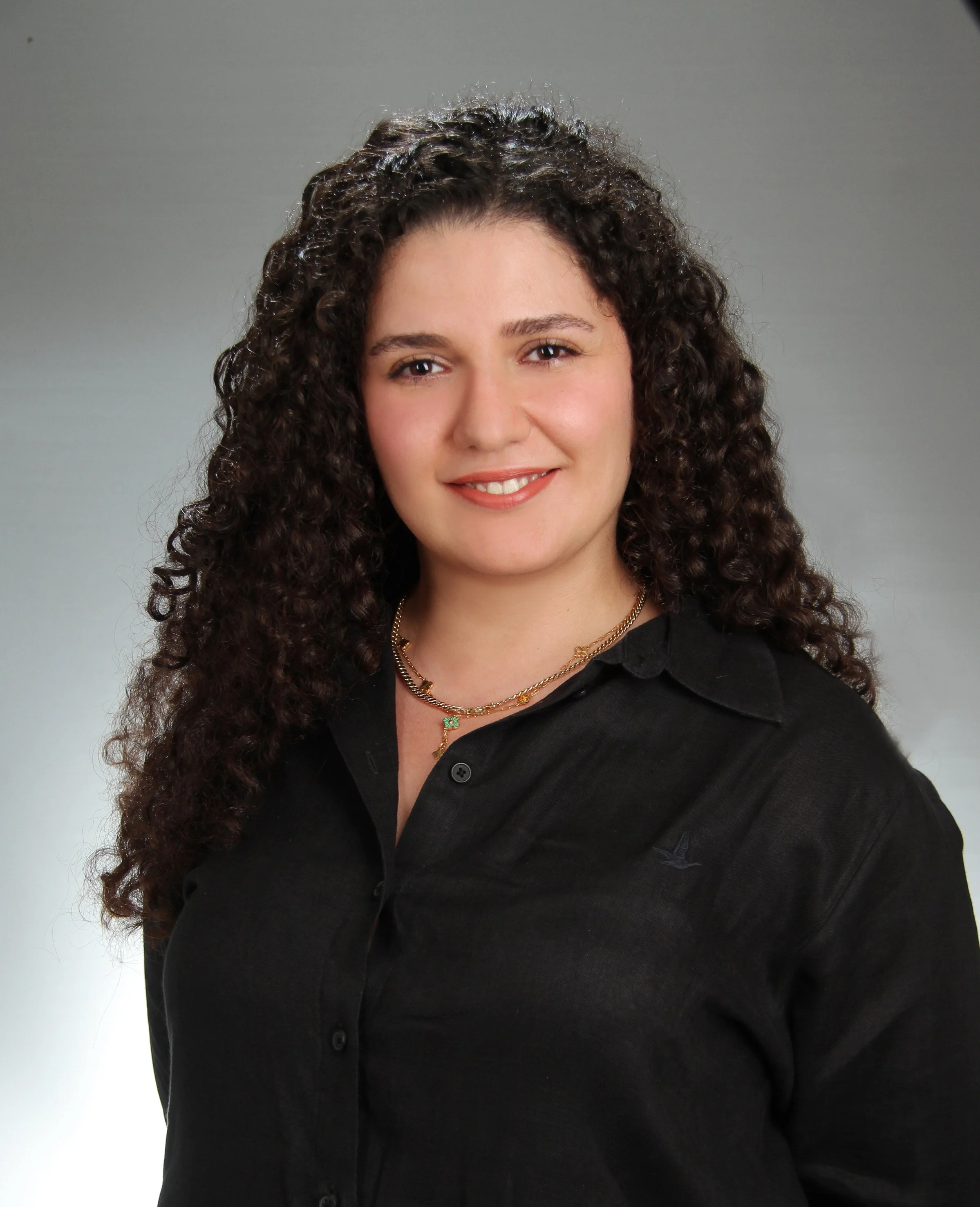 Close-up of a woman with curly brown hair, wearing a black top and layered gold necklaces, smiling softly against a plain gray background.