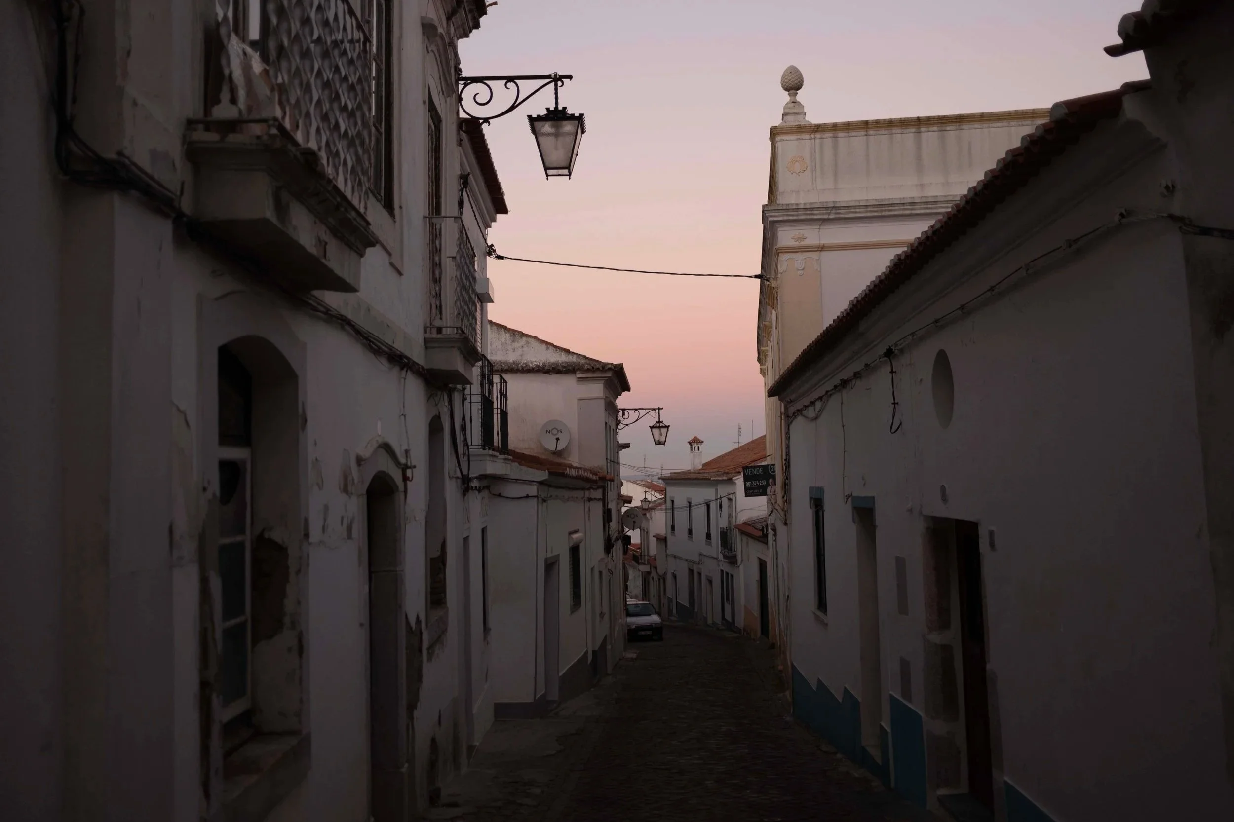 A narrow street lined with white buildings, some with balconies and air conditioning units, at dusk with a pastel purple sky. Street lamps hang from the buildings, and rooftops have terracotta tiles.