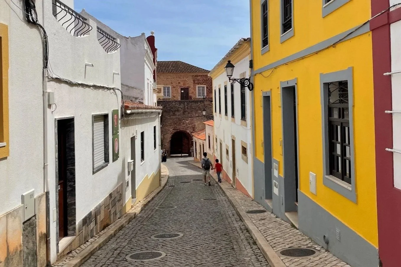 A cobblestone street lined with colorful buildings on a sunny day, with two children and an adult walking toward an arched passageway.