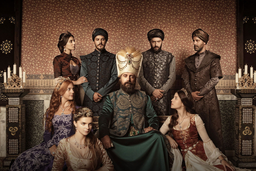 A group of nine people in regal, historical attire posed in a palace setting with ornate decor and candles, suggesting a royal or noble gathering.