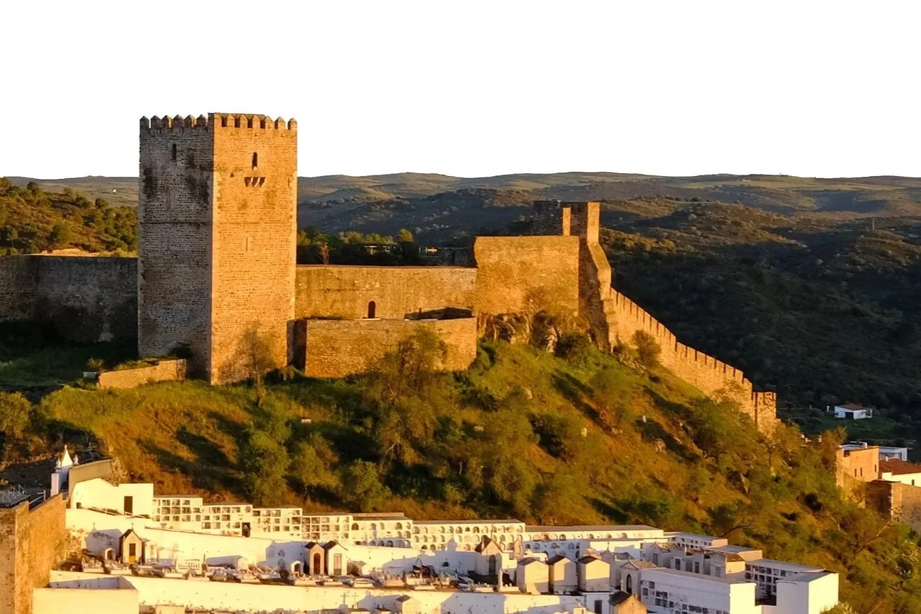 Historic castle with a large tower on a hillside overlooking white buildings in a town, during sunset.